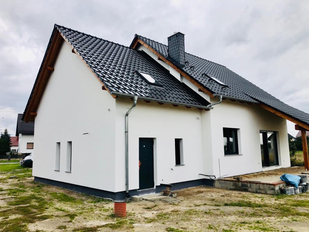 Nowo wybudowany dom jednorodzinny z białą elewacją, czarną dachówką i elementami drewnianymi, widoczne orynnowanie i fragment tarasu w budowie.