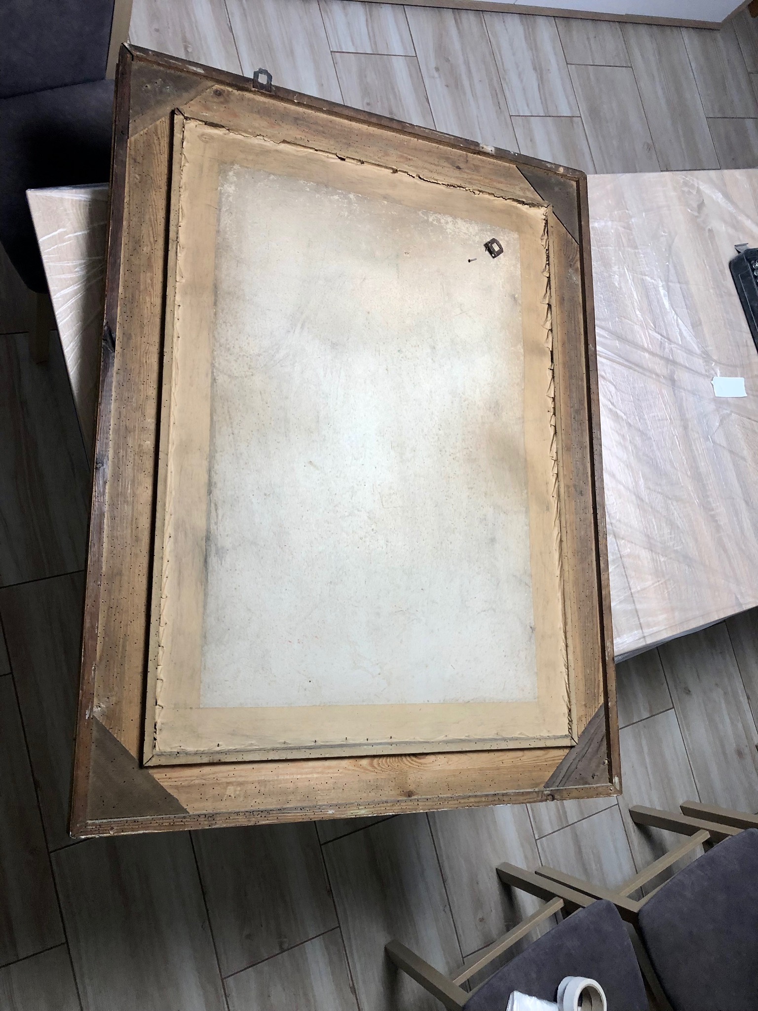 Tył starego obrazu w drewnianej ramie z widocznymi śladami po kornikach, leżący na podłodze obok krzeseł i stolika zabezpieczonego folią.
