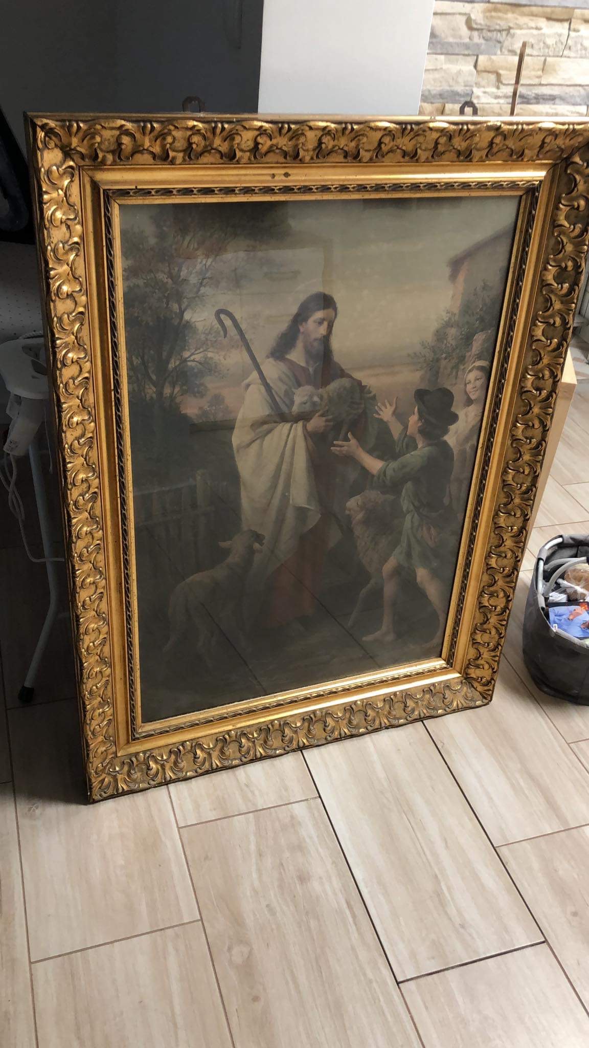 Oprawiony w zdobną, złotą ramę obraz religijny przedstawiający Jezusa z owcami i dziećmi na tle wiejskiego krajobrazu.