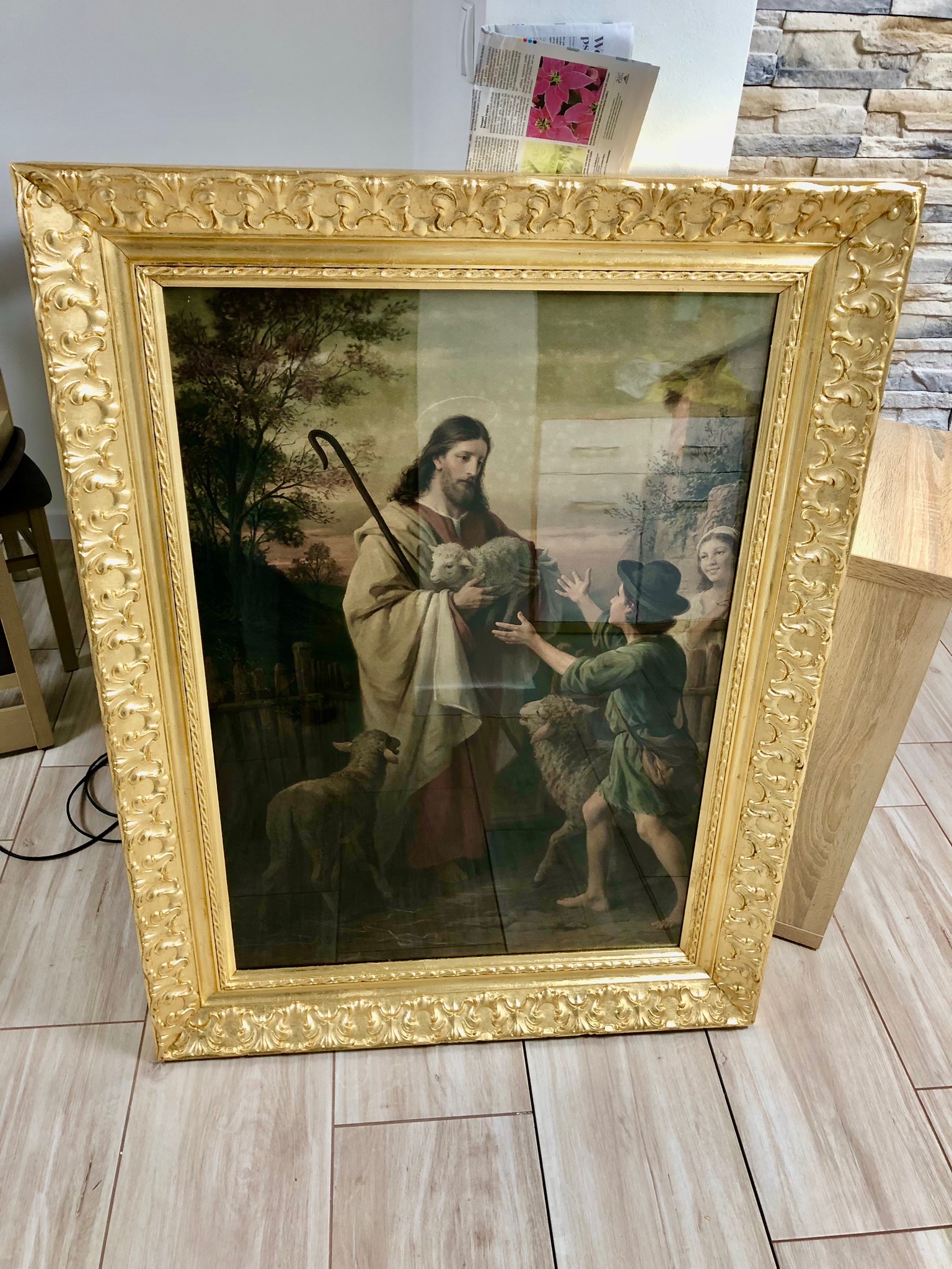 Oprawiony w bogato zdobioną, złotą ramę obraz religijny przedstawiający Jezusa jako dobrego pasterza z owcami i dziećmi na tle pejzażu.