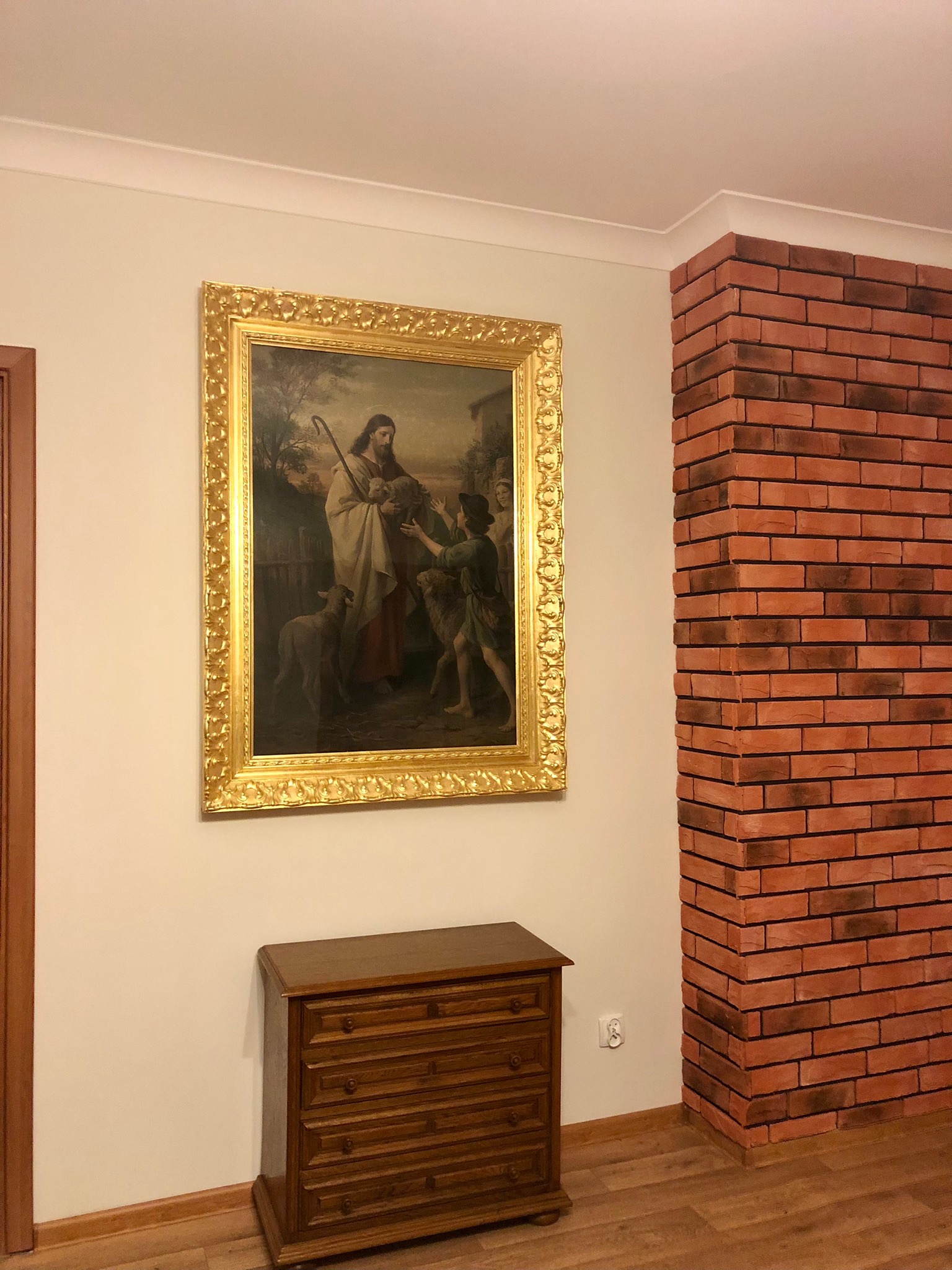 Wnętrze z obrazem w złotej ramie przedstawiającym scenę religijną, komodą i dekoracyjną ścianą z cegły.