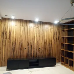 Drewniana ściana z forniru o egzotycznym wzorze, czarna szafka RTV i regał z otwartymi półkami w pasującym fornirze, okrągła lampa sufitowa, widoczne gniazdka elektryczne w ścianie.