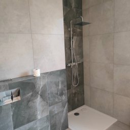 Usługi Kafelkarskie - Nowoczesna łazienka z kabiną prysznicową typu walk-in, wykończona szarymi płytkami imitującymi marmur. Widoczny fragment toalety i rolka papieru.