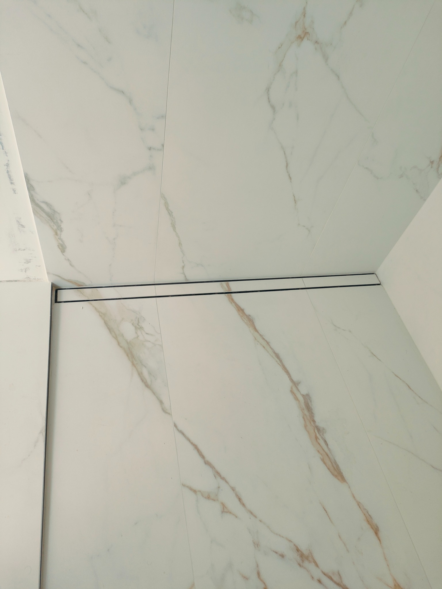 Białe płytki imitujące marmur z odpływem liniowym w kabinie prysznicowej. Minimalistyczny design, precyzyjne ułożenie płytek, widoczny detal odpływu.