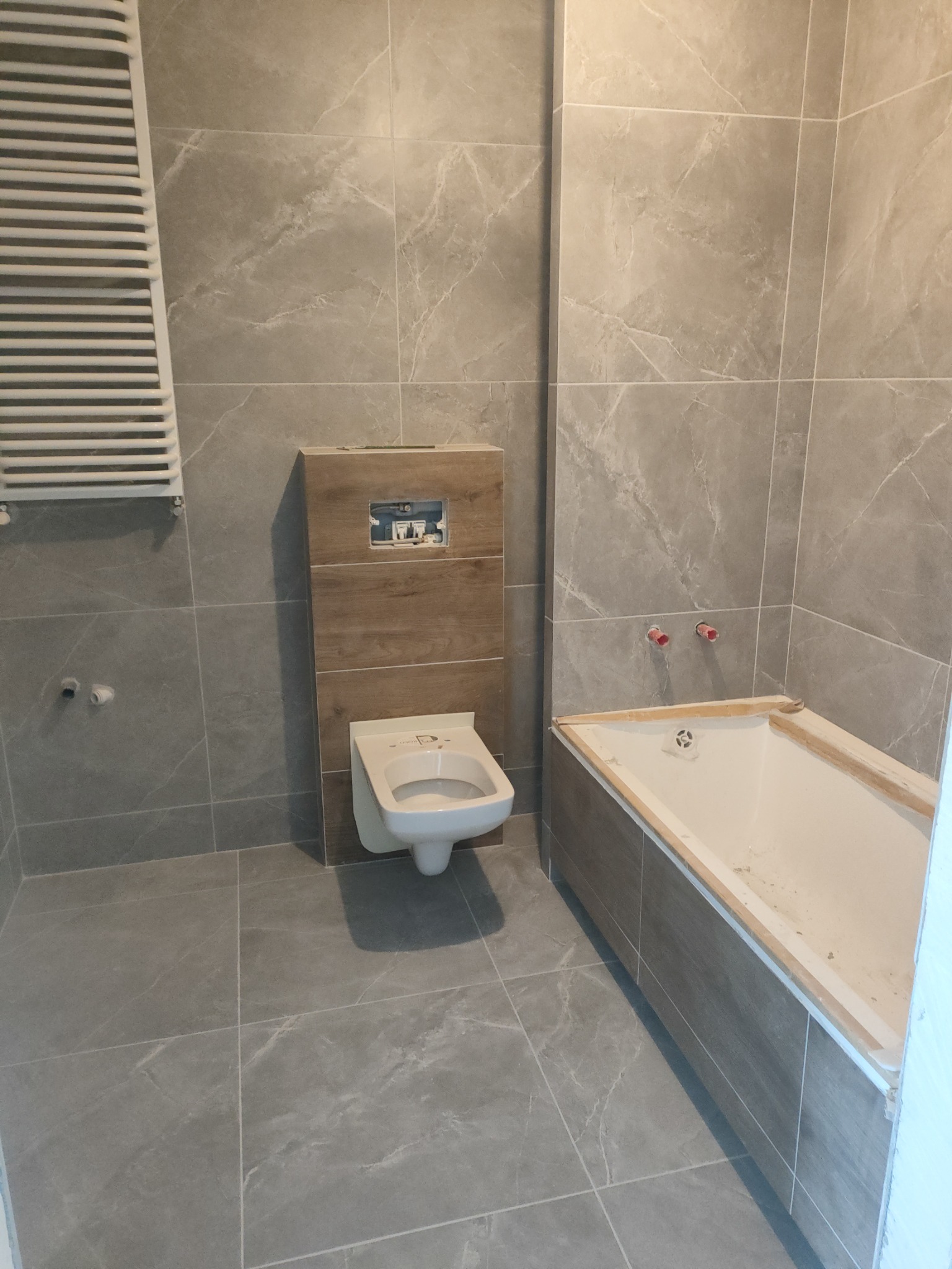 Wykończona łazienka w szarościach z elementami drewna: płytki imitujące marmur na ścianach i podłodze, podwieszana toaleta, zabudowana wanna i grzejnik drabinkowy.