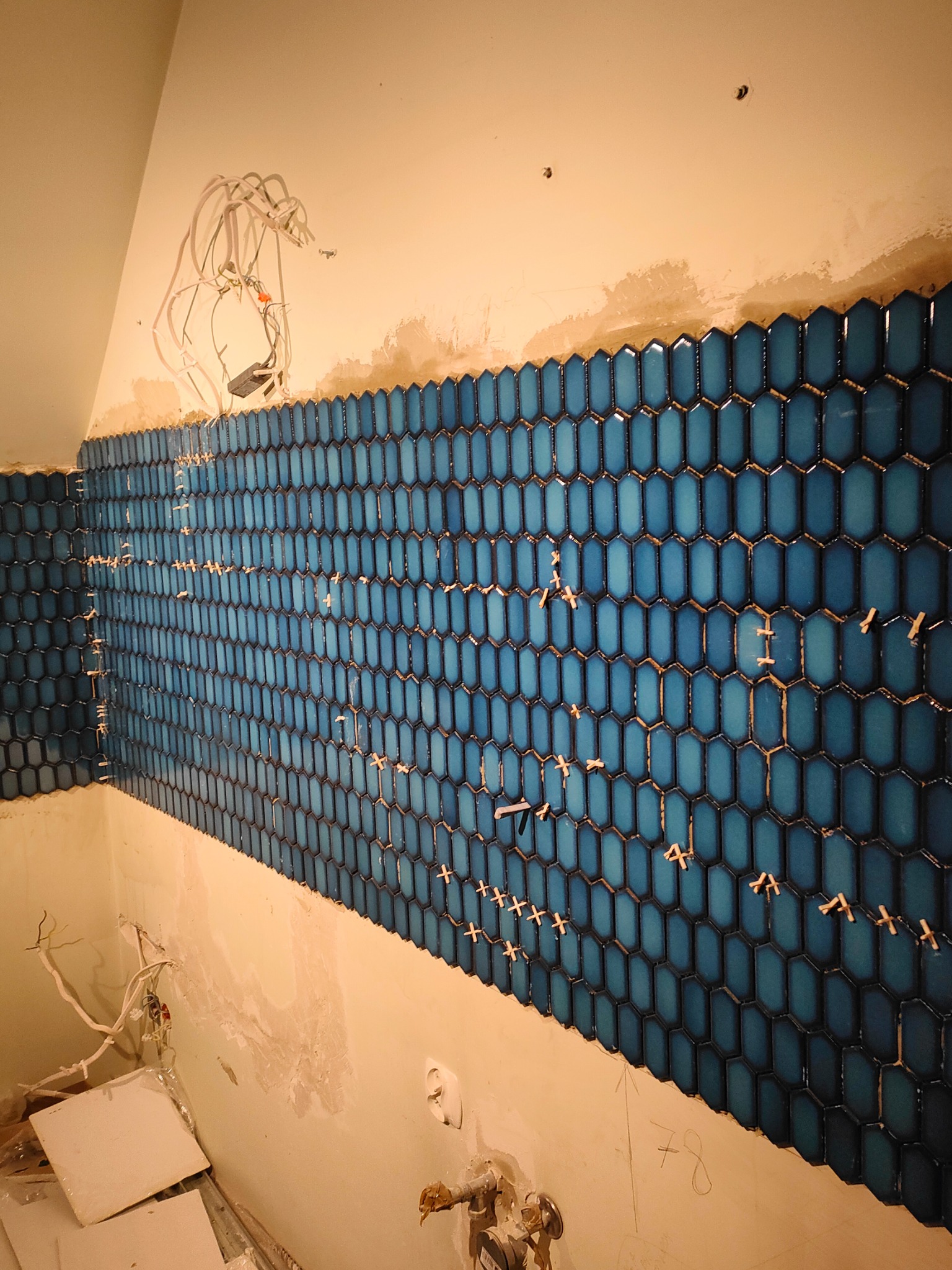 Niebieska mozaika ułożona na ścianie z krzyżykami dystansowymi, widoczne przewody elektryczne i przygotowane podłoże. Nietypowy kształt płytek.