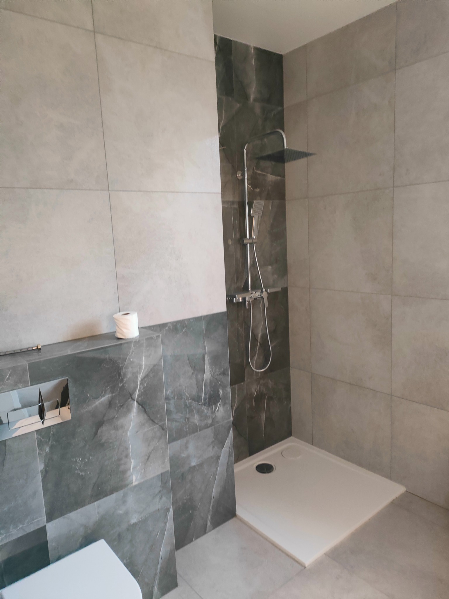 Nowoczesna łazienka z kabiną prysznicową typu walk-in, wykończona szarymi płytkami imitującymi marmur. Widoczny fragment toalety i rolka papieru.