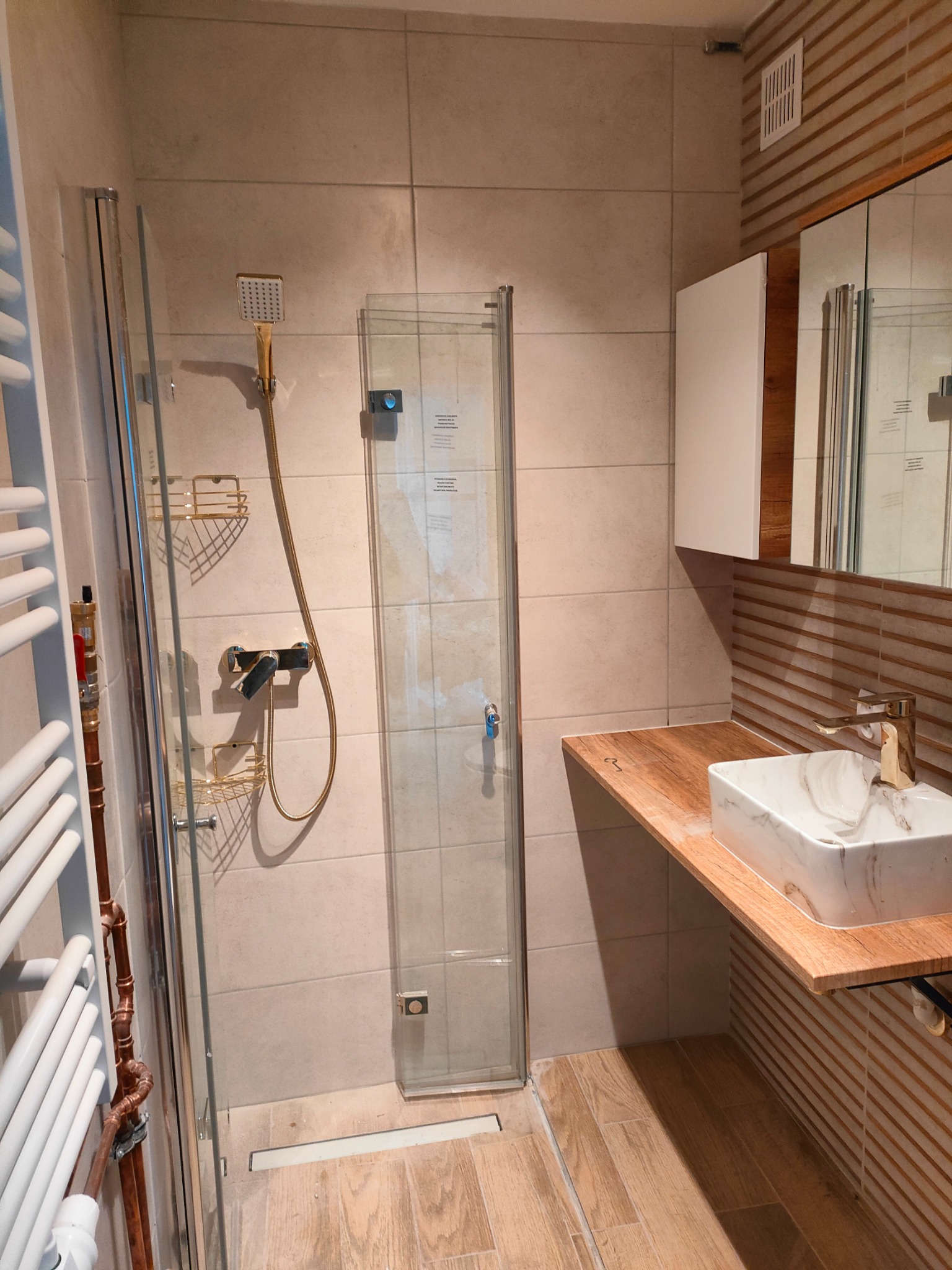 Jasna łazienka po modernizacji: kabina prysznicowa ze złotymi akcentami, umywalka marmurkowa na drewnianym blacie, grzejnik drabinkowy i beżowe płytki.