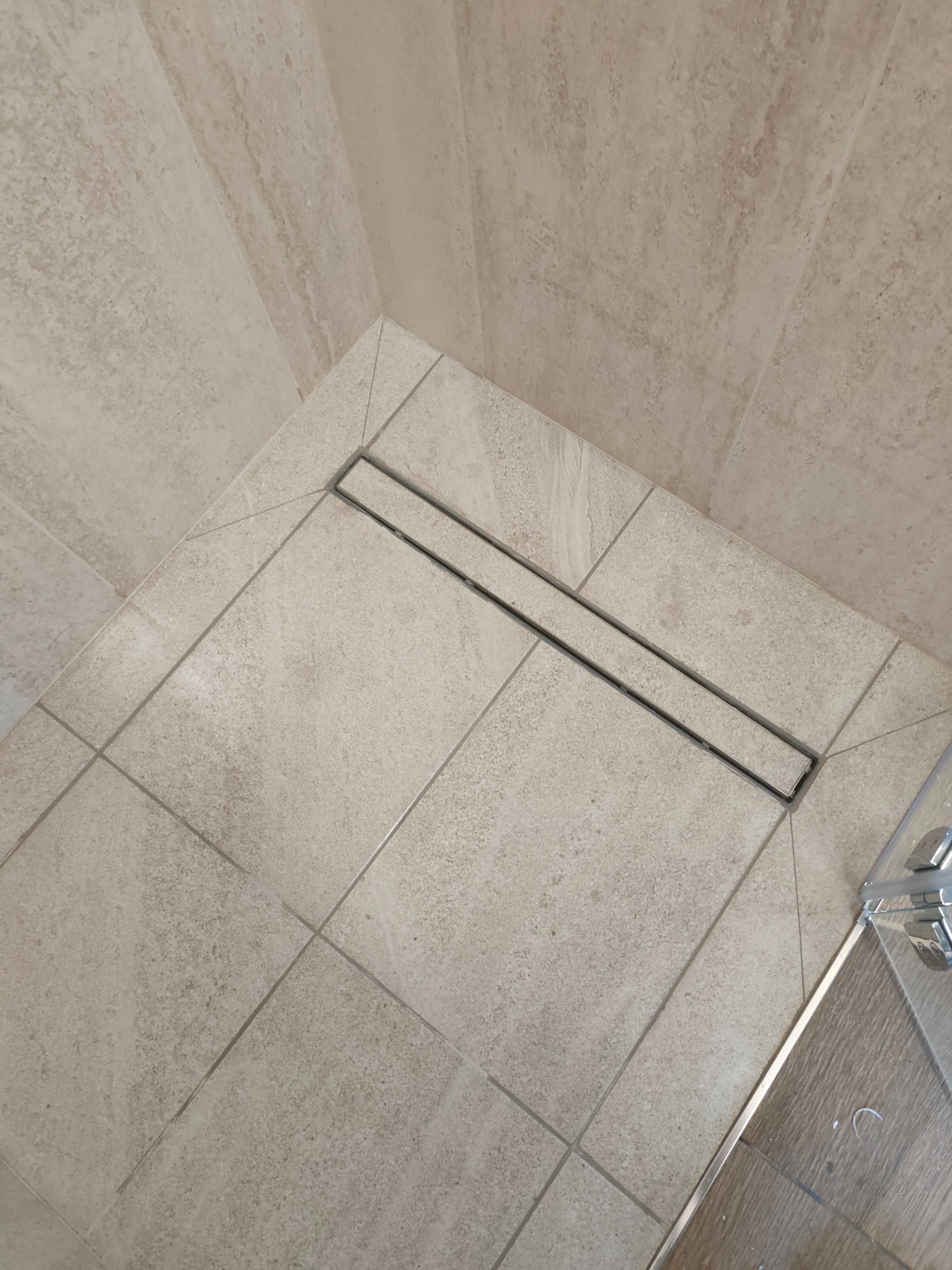 Beżowe płytki podłogowe z odpływem liniowym w kabinie prysznicowej, widok z góry. Minimalistyczny design, precyzyjne fugi, fragment szklanej ścianki kabiny.