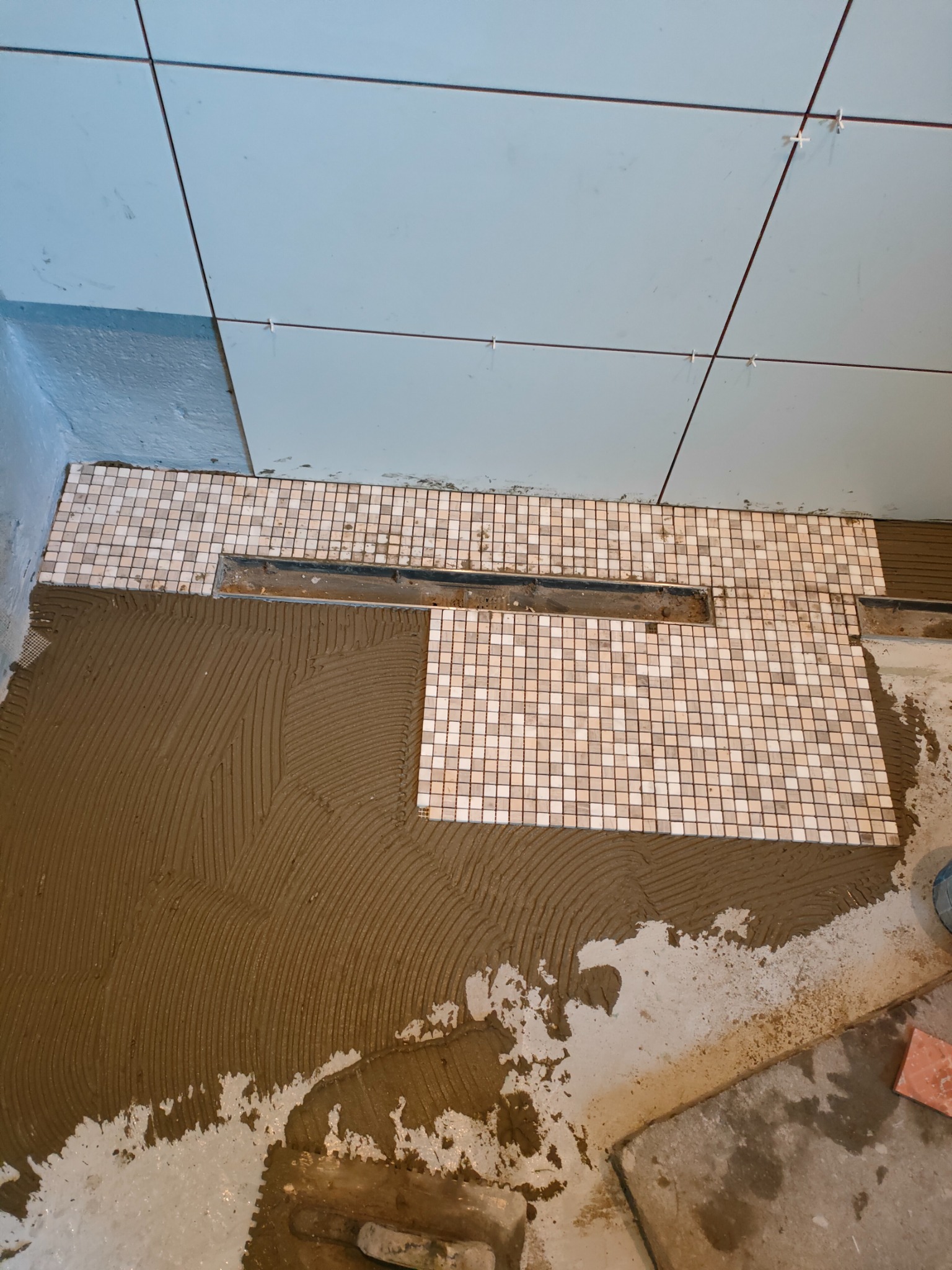 Układanie mozaiki w łazience, widoczny odpływ liniowy i świeżo nałożony klej. Jasne płytki na ścianie z krzyżykami dystansowymi.