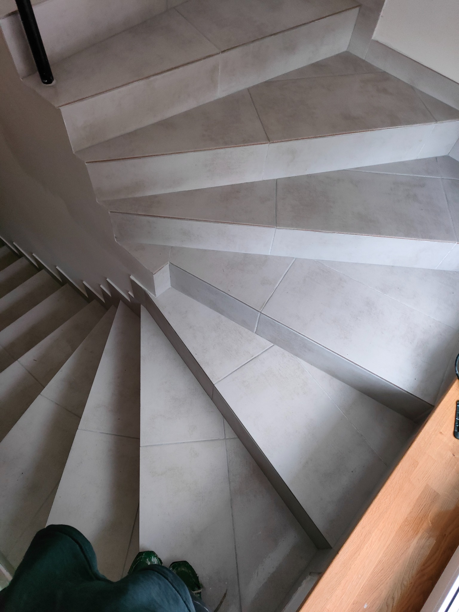 Spiralne schody wyłożone jasnymi, kwadratowymi płytkami, widok z góry. Fragment osoby w zielonym ubraniu w dolnej części kadru. Poręcz schodowa po lewej stronie.