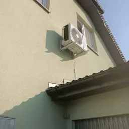 Biały klimatyzator zamontowany na elewacji domu, widoczne przewody i wsporniki, zarys okna i fragment dachu.