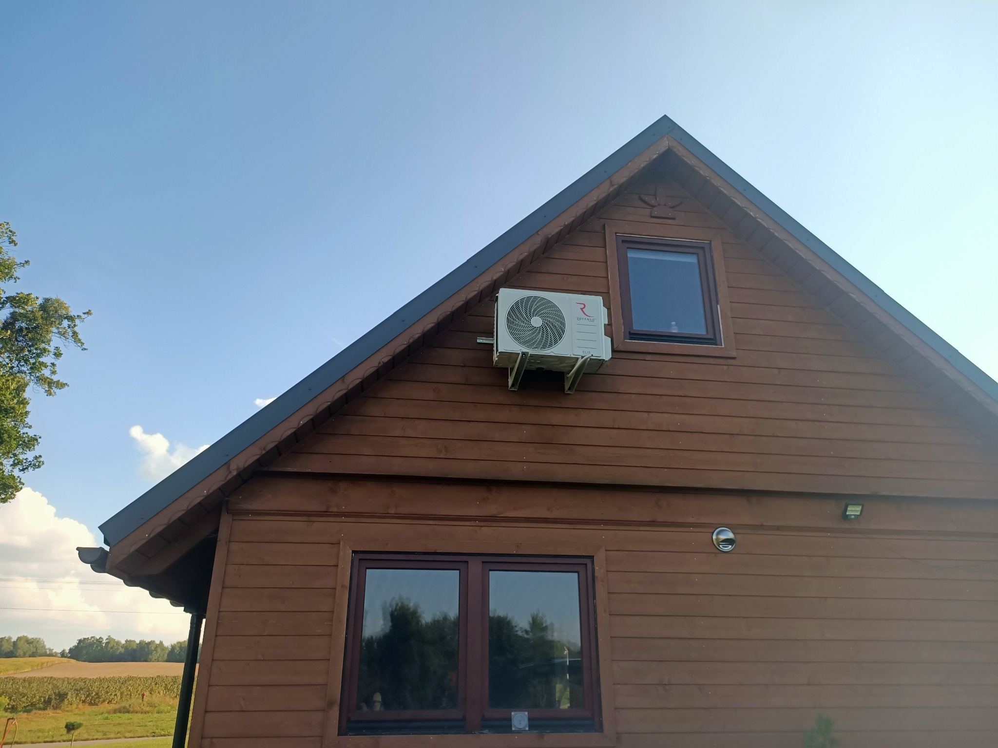 Biała jednostka zewnętrzna klimatyzacji marki Rotenso zamontowana na brązowej, drewnianej elewacji domu jednorodzinnego z dwoma oknami, na tle błękitnego nieba.