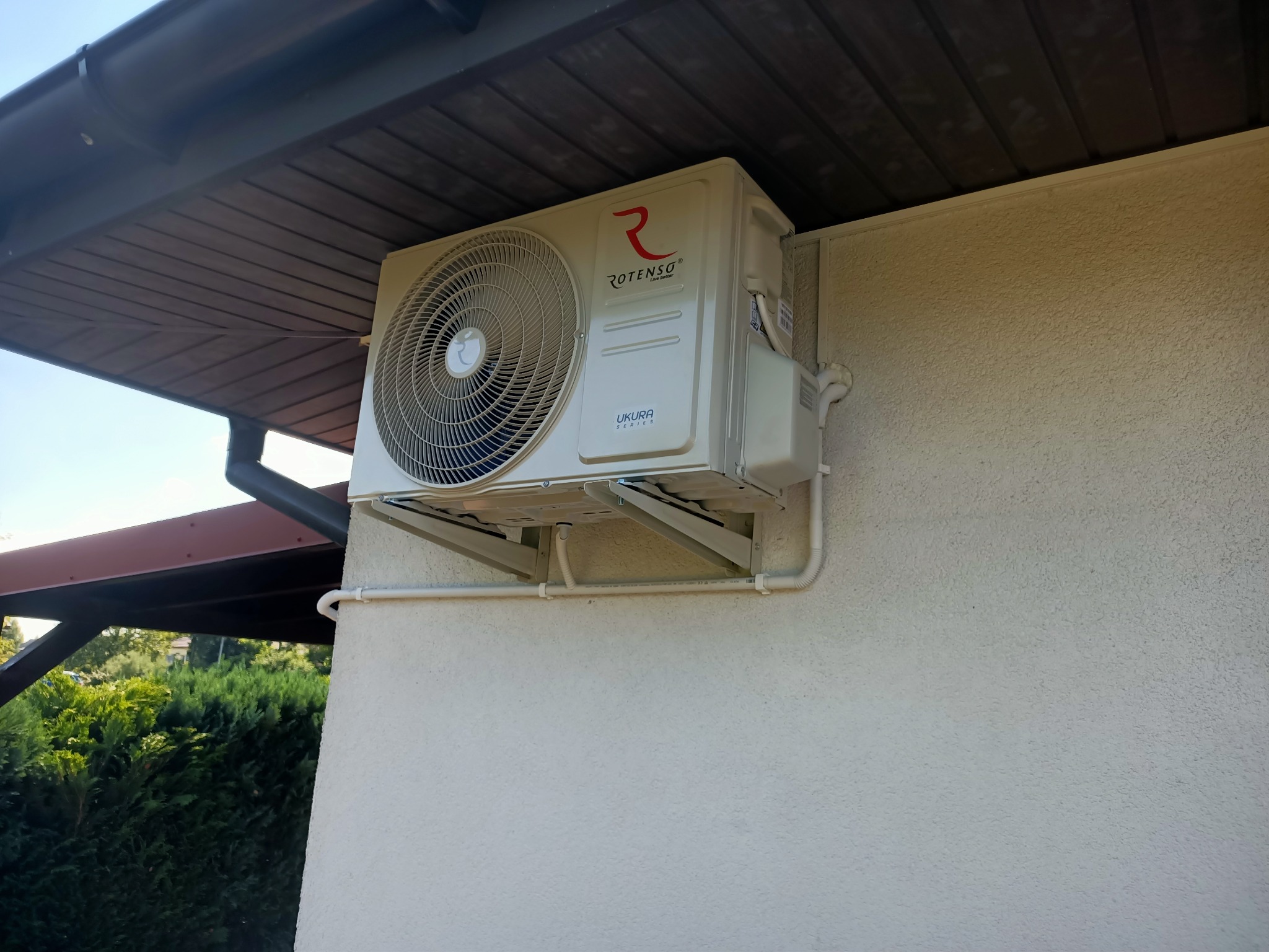 Zamontowana jednostka zewnętrzna klimatyzacji Rotenso Ukura Series z widocznymi rurami instalacyjnymi na elewacji budynku z fakturą baranka, zadaszona konstrukcją drewnianą.