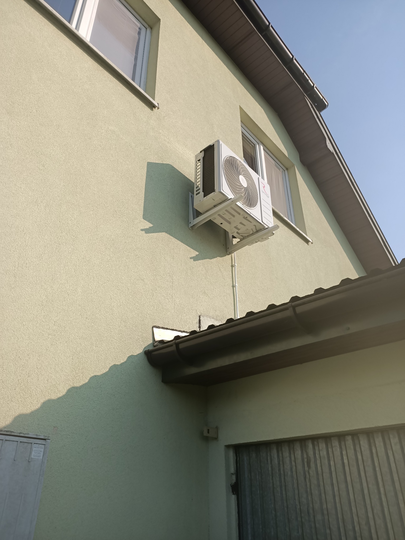 Biały klimatyzator zamontowany na elewacji domu, widoczne przewody i wsporniki, zarys okna i fragment dachu.