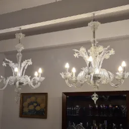 Dwa kryształowe żyrandole w stylu weneckim z żarówkami świecowymi, jeden nad obrazem z motywem kwiatowym, drugi nad przeszkloną witryną z kieliszkami.