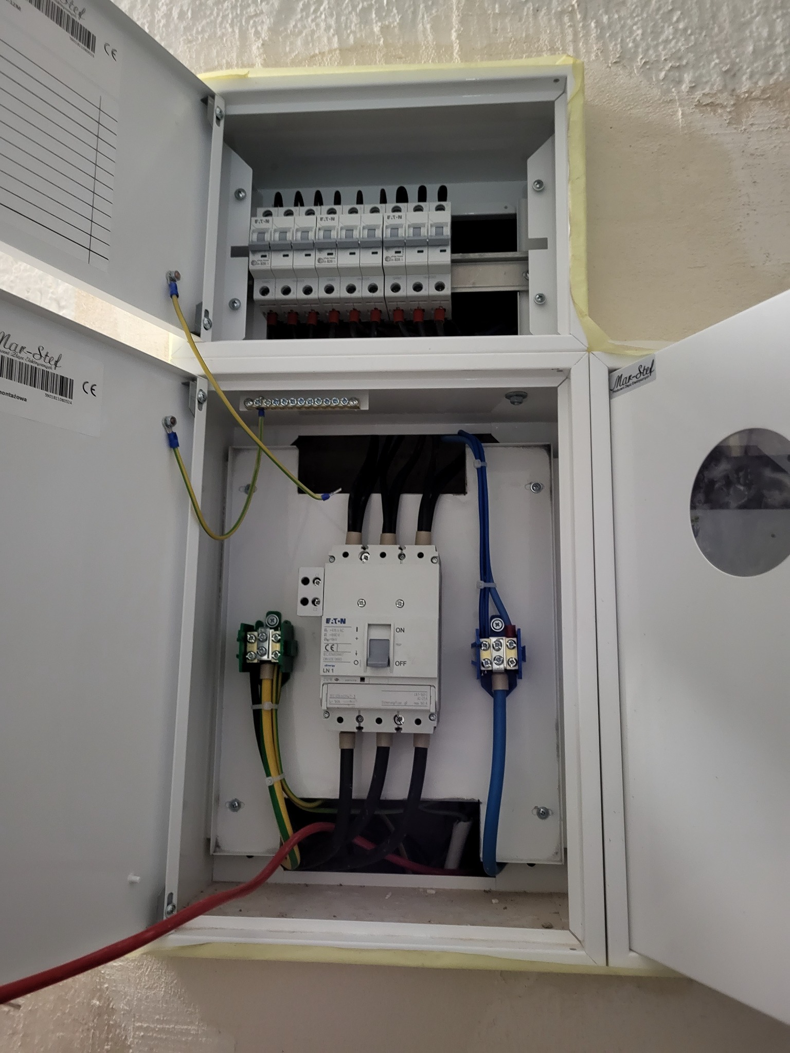 Otwarta skrzynka elektryczna z zainstalowanymi bezpiecznikami, przewodami i aparaturą modułową, widoczne oznaczenia producenta Eaton oraz naklejka Mar-Stel na drzwiczkach.