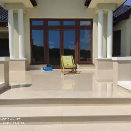 Jasne, polerowane płytki na schodach i tarasie przed domem z białymi kolumnami i brązowymi oknami. Na tarasie stoi zielone krzesło plażowe.