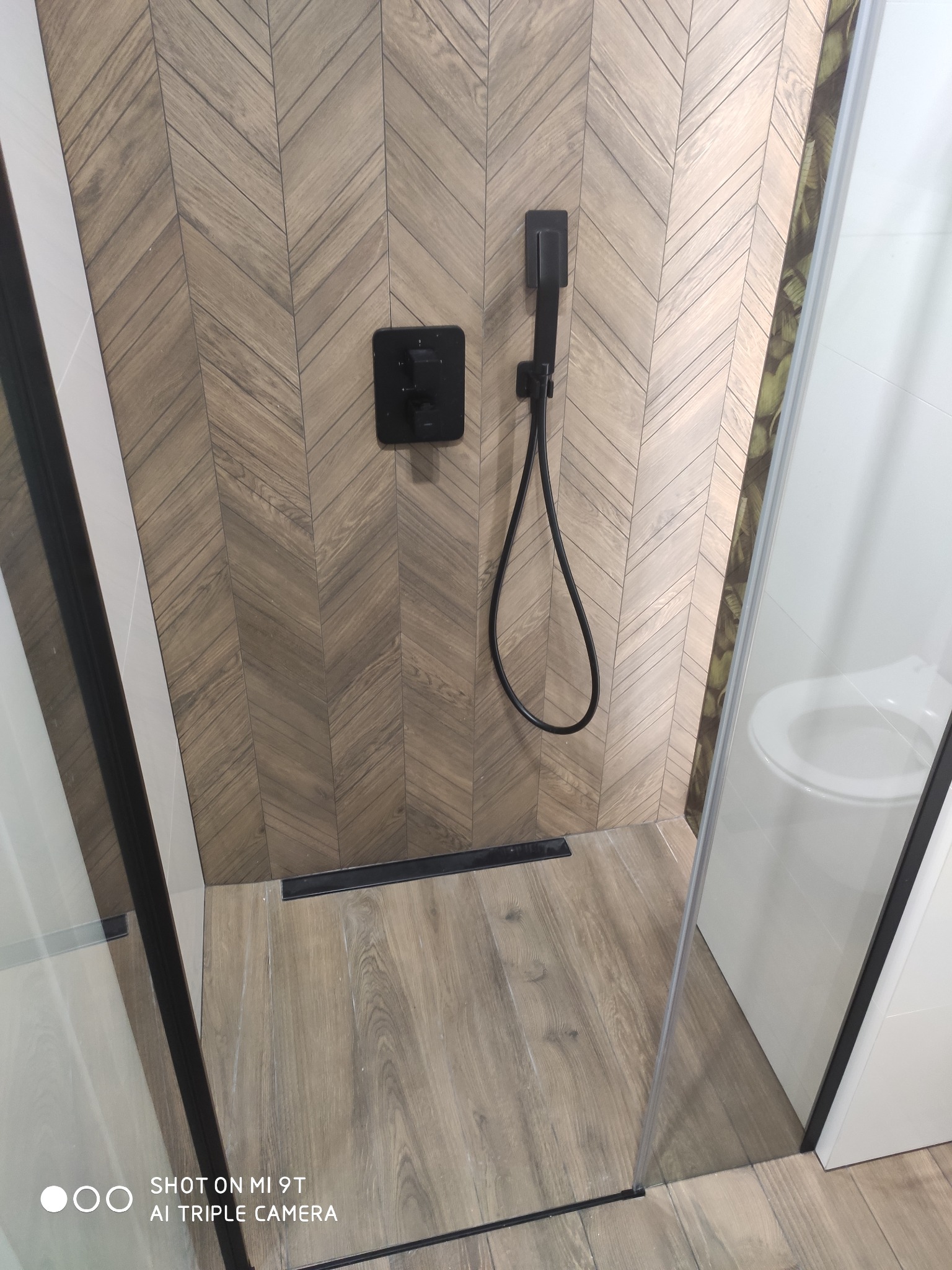 Kabina prysznicowa z drewnopodobnymi płytkami ułożonymi w jodełkę, czarna armatura i odpływ liniowy, widoczny fragment toalety za szklaną ścianką.