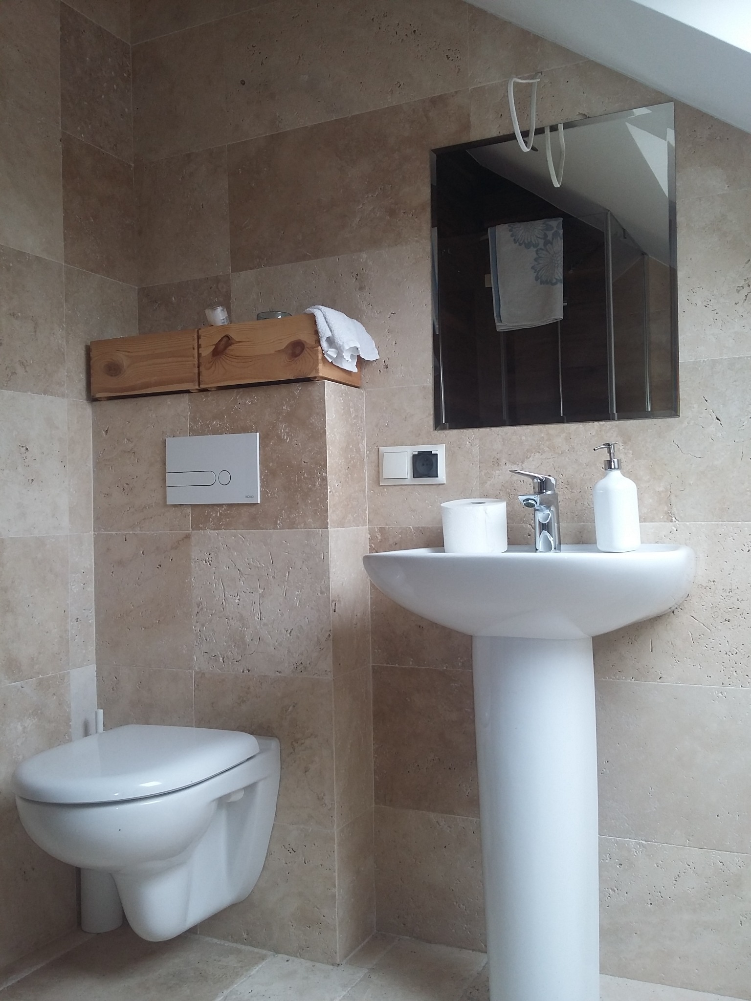 Nowoczesna łazienka z beżowymi płytkami trawertyn, wisząca toaleta, umywalka na postumencie, drewniana półka z kosmetykami i lustro