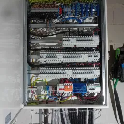 Otwarta skrzynka rozdzielcza z widocznymi bezpiecznikami, przewodami w różnych kolorach i złączkami, zamontowana na ścianie z surowej cegły.