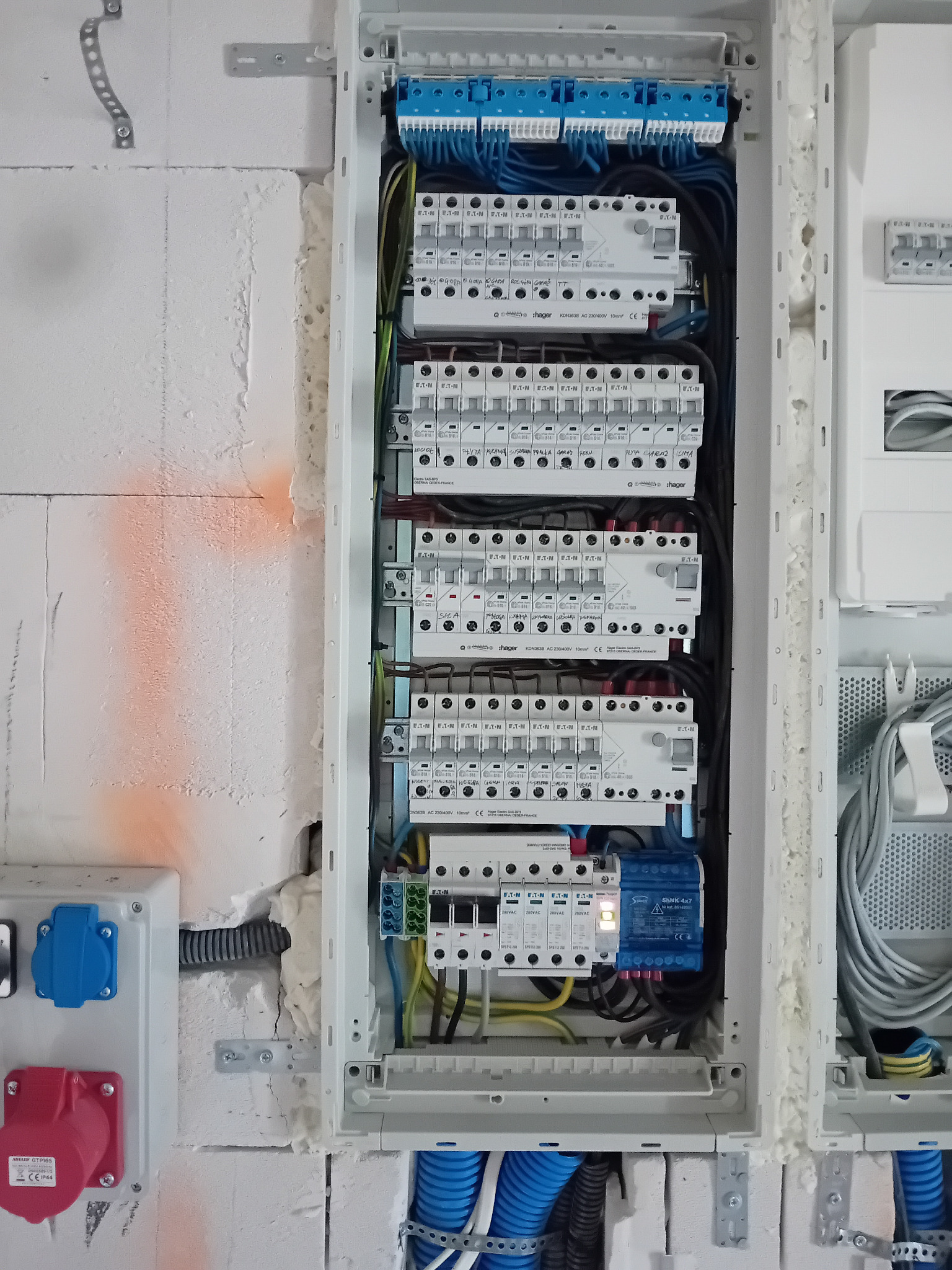 Rozdzielnica elektryczna z bezpiecznikami i przewodami w białej obudowie, zamontowana na ścianie z betonu komórkowego z widocznymi śladami pianki montażowej i pomarańczowym oznaczeniem, z gniazdami...
