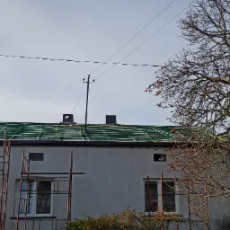 W trakcie krycia dachu zieloną blachodachówką, widoczne rusztowanie przy ścianie budynku z oknami, drzewo z brązowymi liśćmi w tle, słupy elektryczne na dachu.