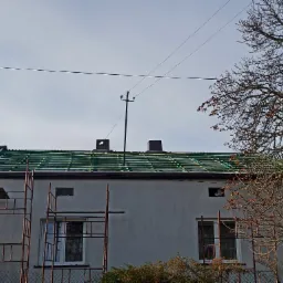 Budynek z widoczną konstrukcją dachu przygotowaną pod pokrycie, rusztowanie przy ścianie, kominy i linia energetyczna na tle jasnego nieba.