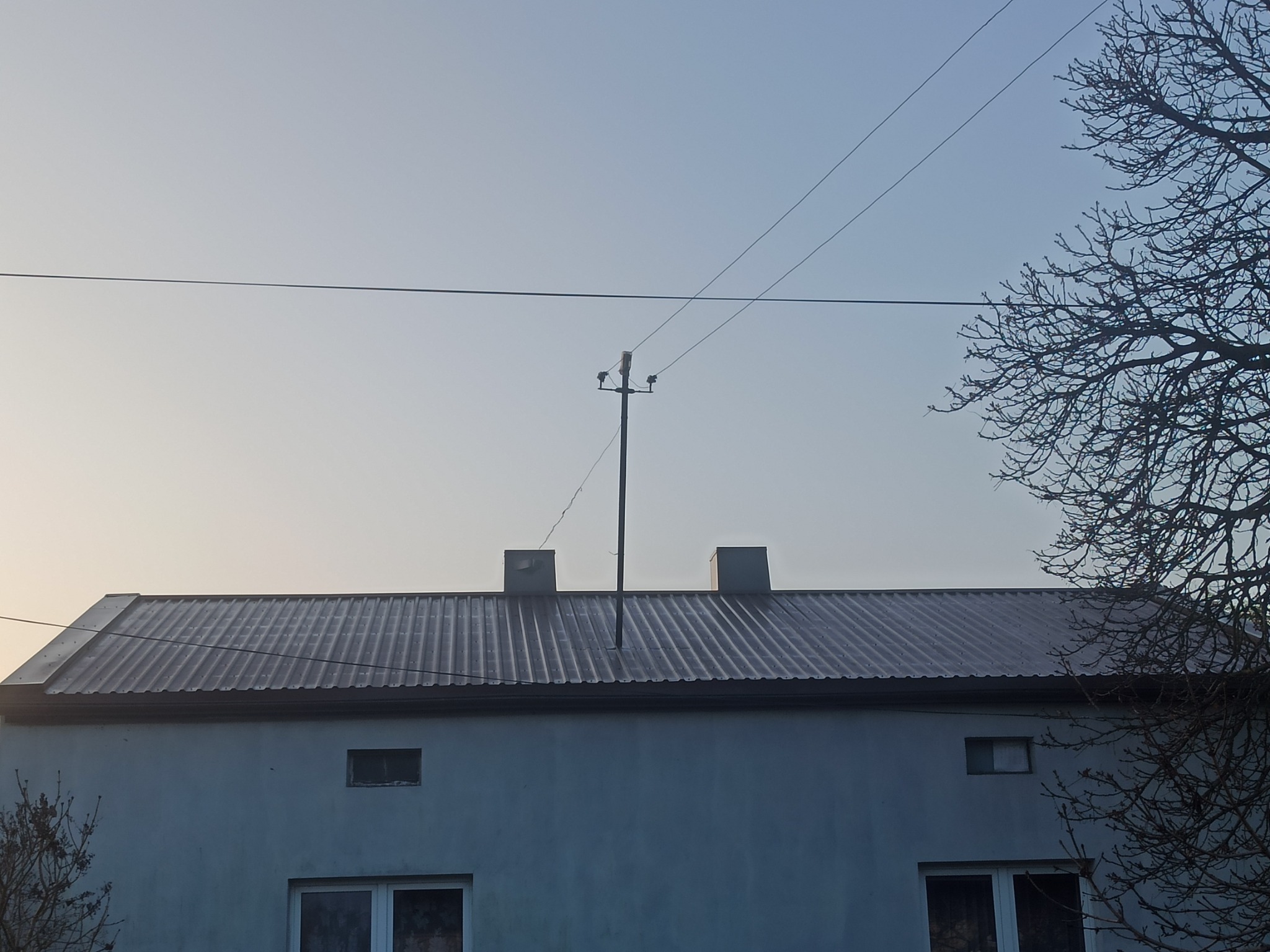 Dom z szarym dachem z blachy falistej, widoczne kominy i słup elektryczny na tle bladego nieba i gałęzi drzewa.