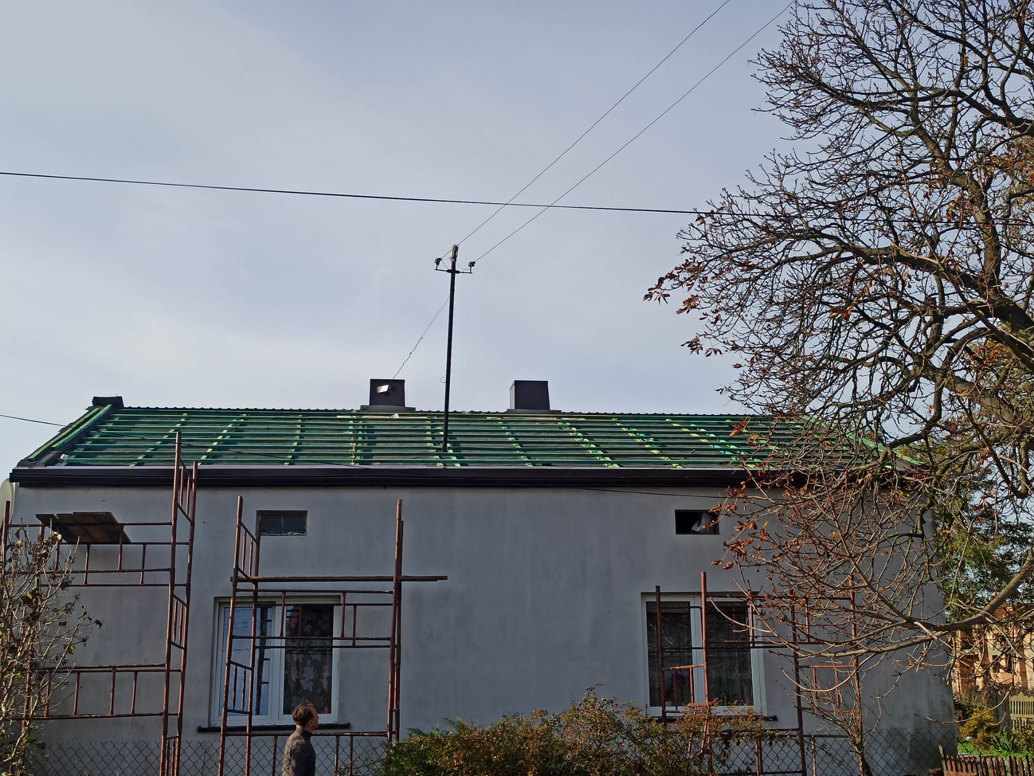 Dom jednorodzinny z widoczną konstrukcją dachu pokrytą zieloną folią, przygotowaną pod dalsze prace dekarskie, rusztowania przy ścianie budynku, jesienne drzewo w tle.