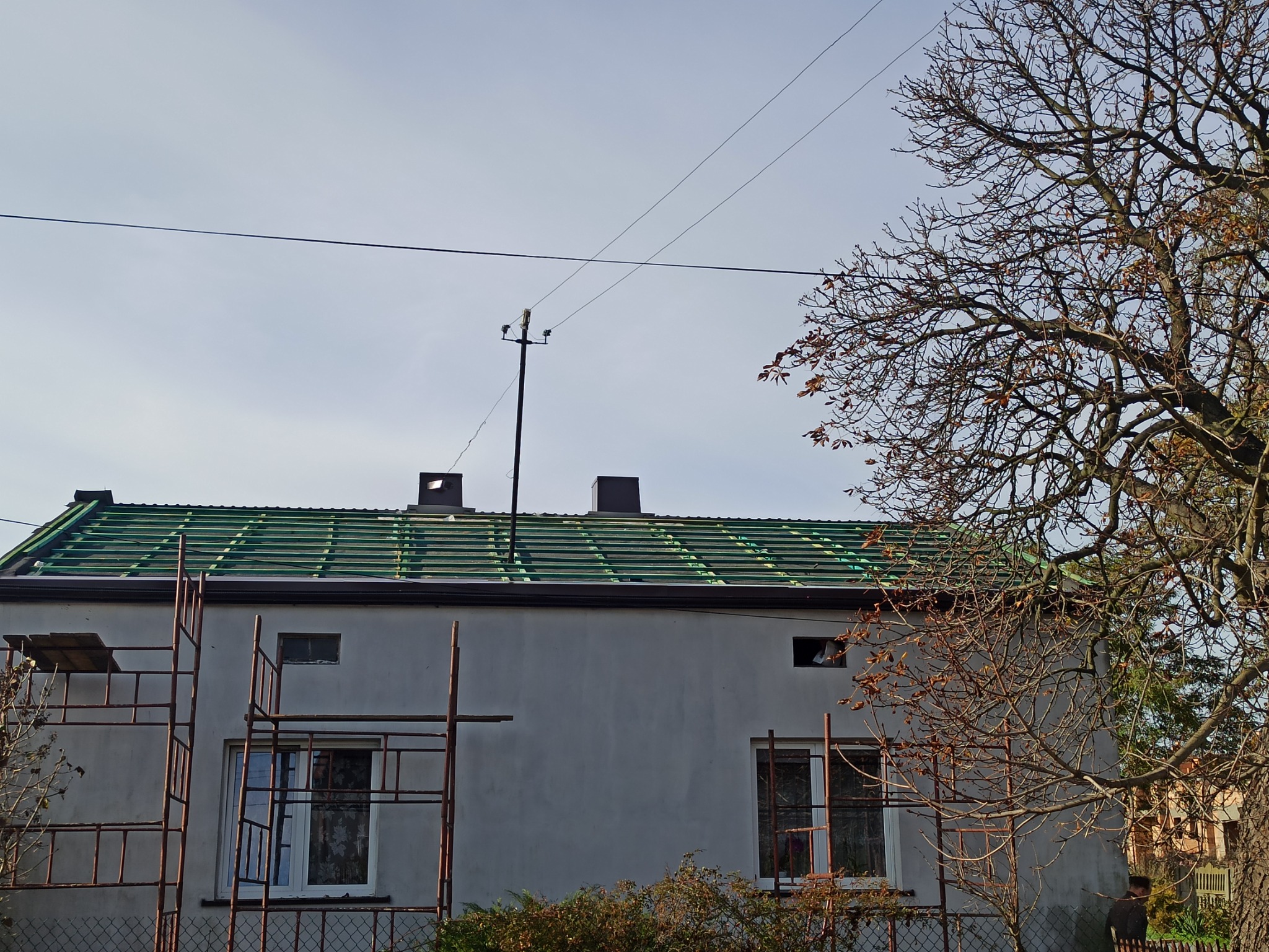 W trakcie krycia dachu zieloną blachodachówką, widoczne rusztowanie przy ścianie budynku z oknami, drzewo z brązowymi liśćmi w tle, słupy elektryczne na dachu.