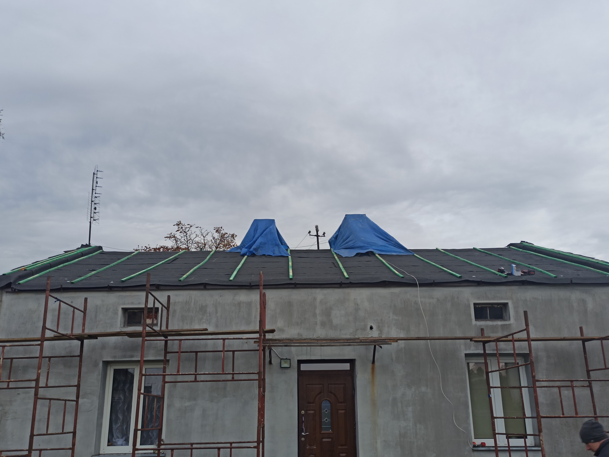 Dom w trakcie prac dekarskich: czarna papa na dachu z zielonymi łatami, zabezpieczona niebieską plandeką, rusztowanie przy ścianie budynku.