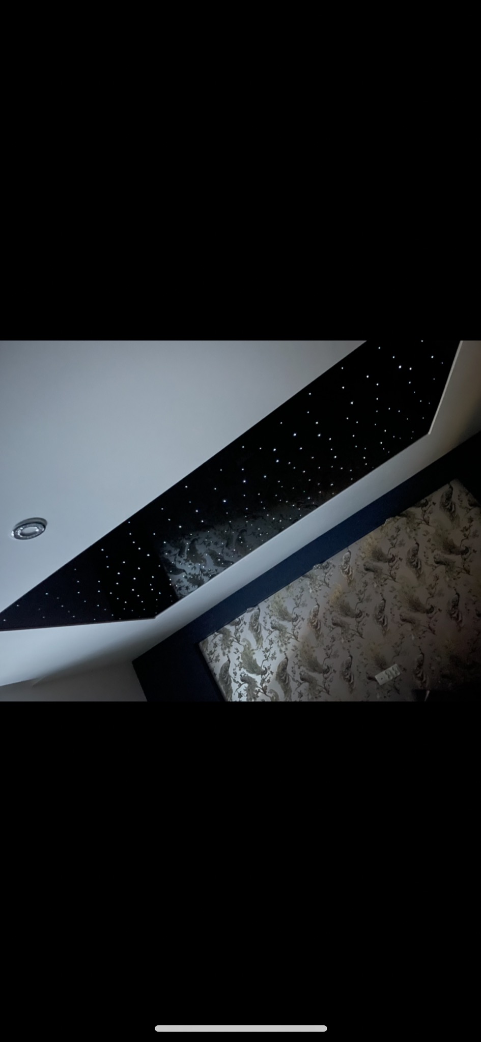 Czarny sufit napinany z efektem gwiazd, oświetlenie punktowe w bieli, ściana z tapetą w odcieniach szarości z motywem pawich piór.