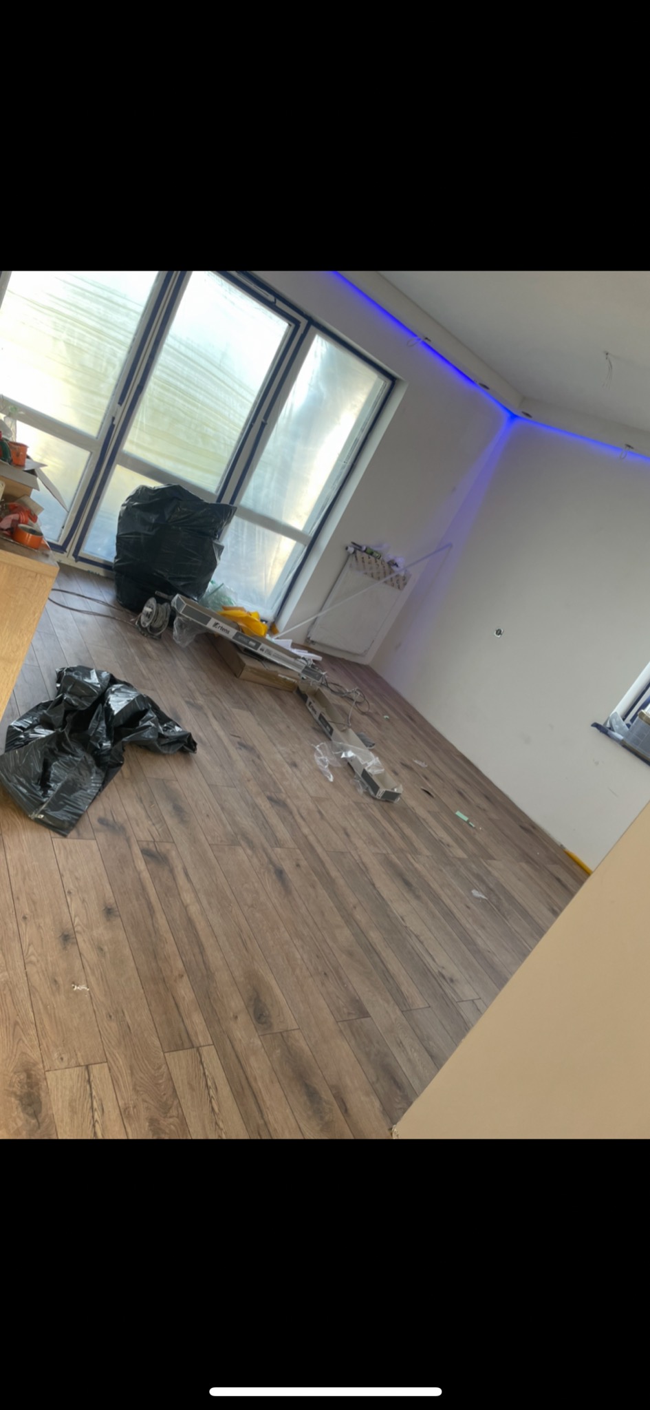 Pokój w trakcie prac wykończeniowych: panele na podłodze, okno zabezpieczone folią, śmieci w workach, listwy przypodłogowe, oświetlenie LED przy suficie.