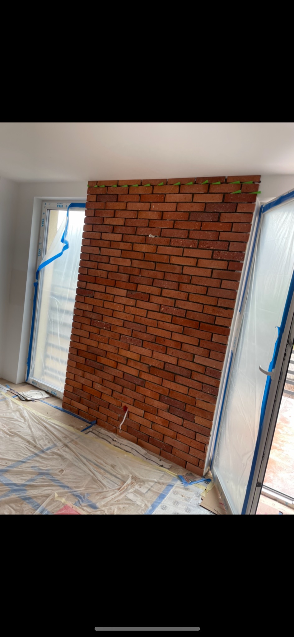 Ściana wyłożona czerwoną cegłą dekoracyjną, zabezpieczone okna folią malarską, widoczne zielone kliny dystansowe. Prace w trakcie realizacji.