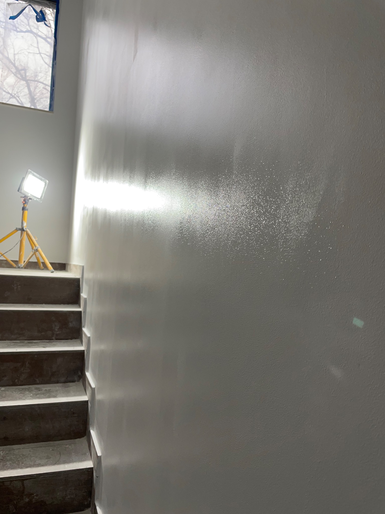 Świeżo pomalowana szara ściana na klatce schodowej, oświetlona lampą budowlaną na trójnogu, widoczne ślady po malowaniu. Okno zabezpieczone folią malarską.