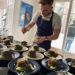 Kucharz w okularach i granatowym fartuchu dekoruje mikro-ziołami talerze z tatarem z tuńczyka na awokado z nori, przygotowane do serwowania.