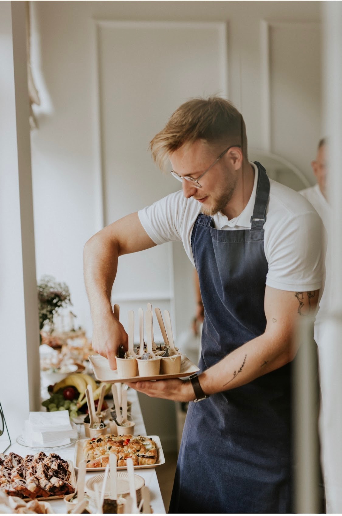 Mężczyzna w okularach i niebieskim fartuchu przygotowuje deserki w papierowych kubeczkach, ustawiając je na tacy z drewna, na stole widoczne inne przekąski.