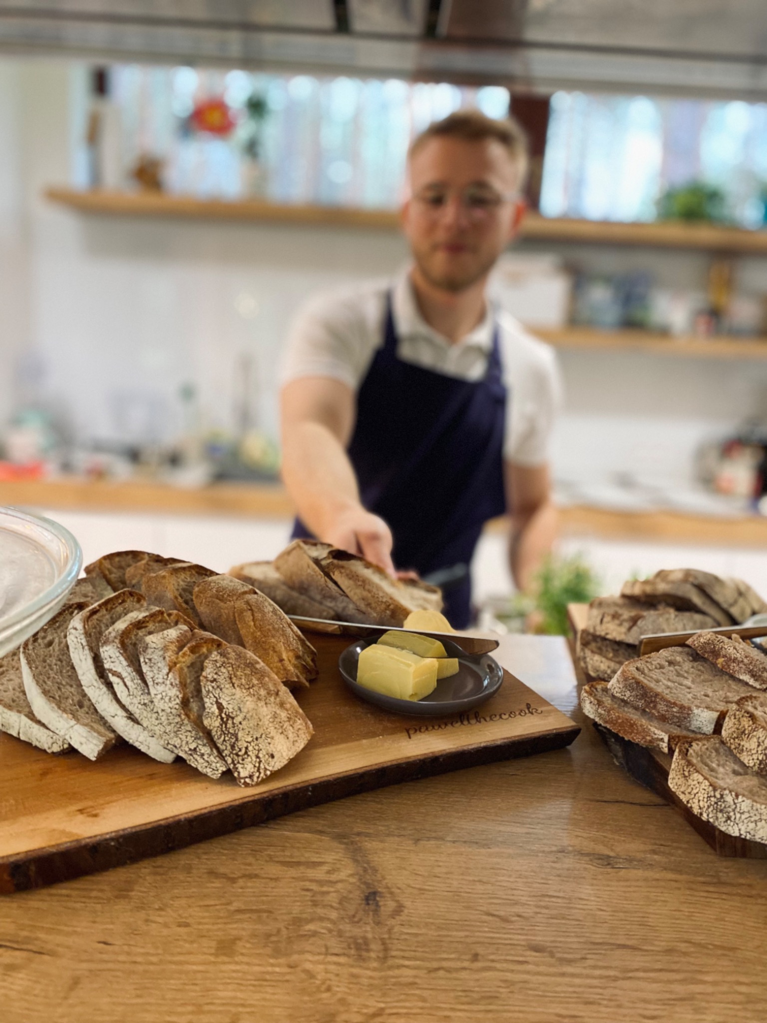 Kucharz w niebieskim fartuchu podaje kromkę chleba z deską serów na drewnianym blacie. Na desce wygrawerowany napis 'pawellthecook'.