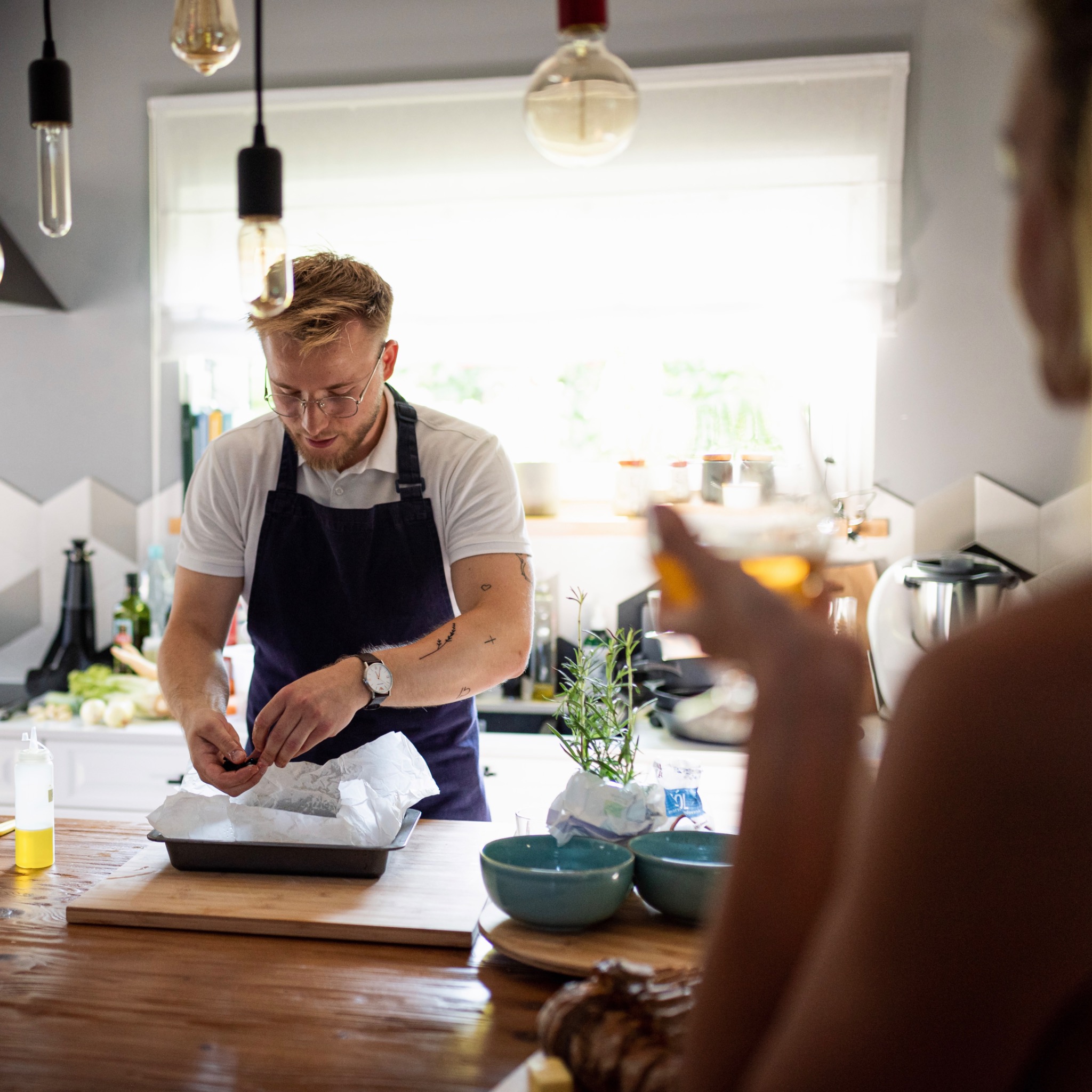 Kucharz w okularach i fartuchu układa oliwki w formie do pieczenia wyłożonej papierem, na drewnianym blacie w jasnej kuchni, z widoczną osobą trzymającą drinka w tle.