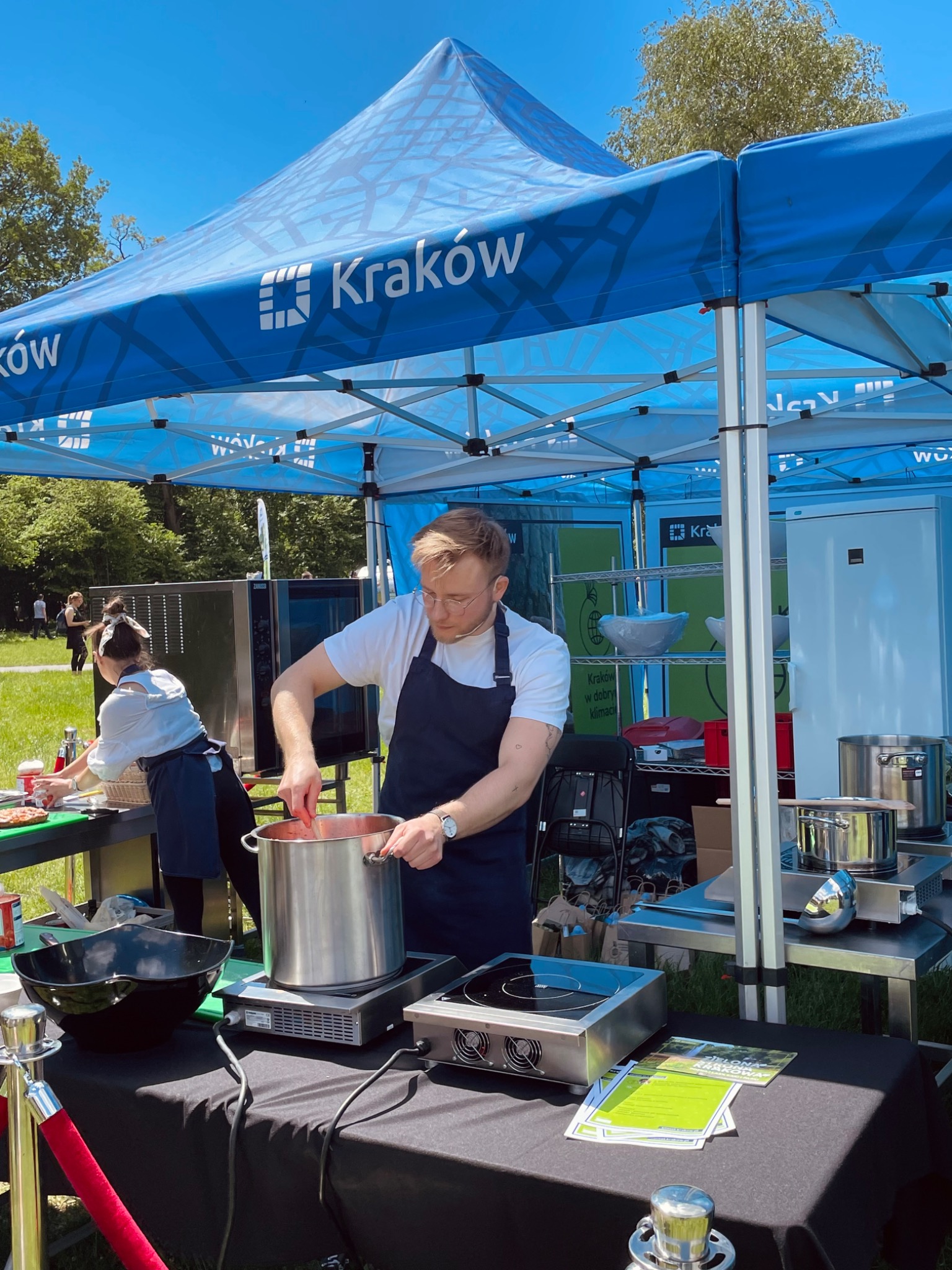 Kucharz miesza zawartość dużego, metalowego garnka na przenośnej kuchence indukcyjnej pod niebieskim namiotem promocyjnym miasta Kraków podczas wydarzenia plenerowego.