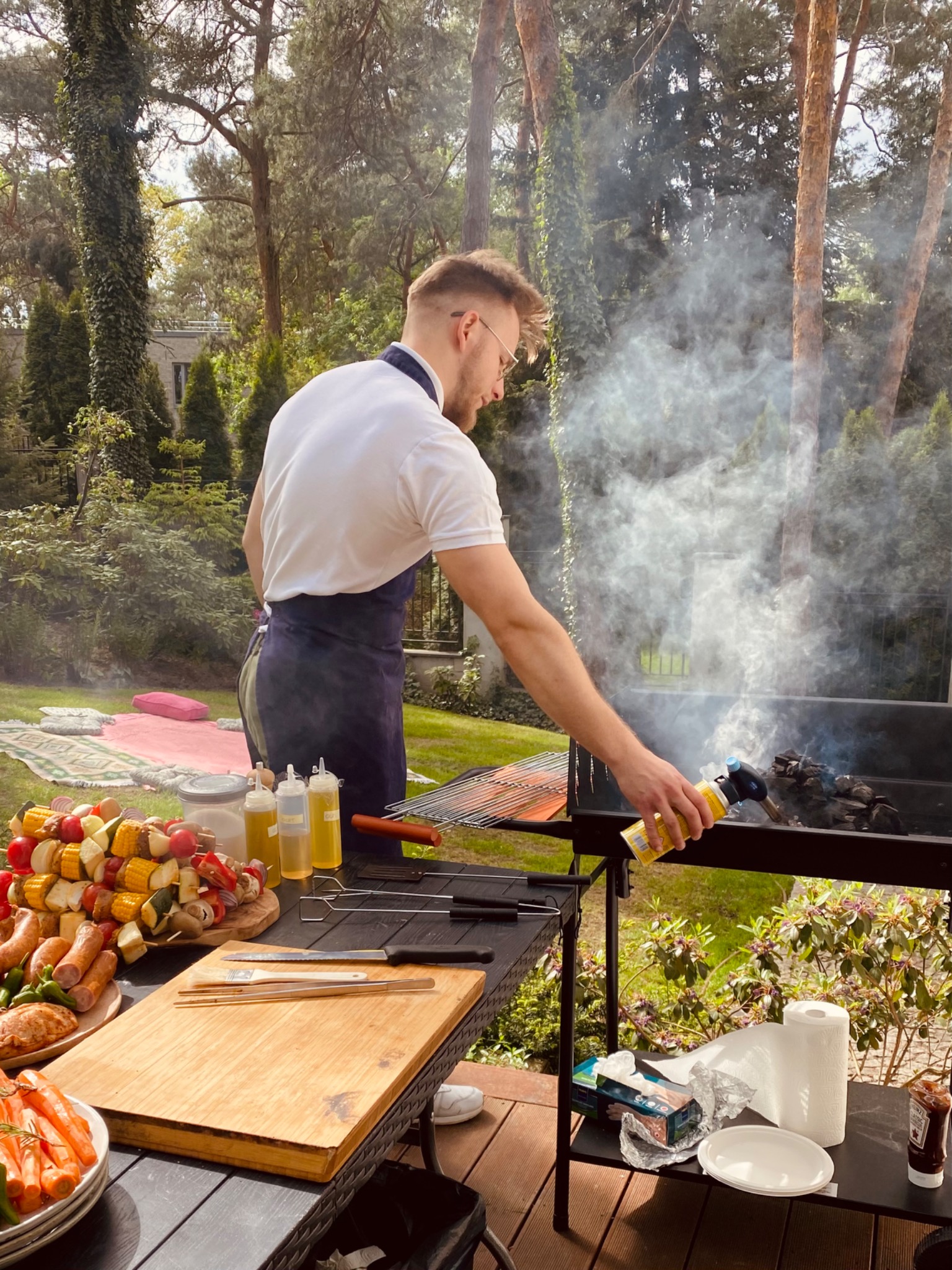 Mężczyzna w fartuchu rozpala grilla na zewnątrz, obok przygotowane szaszłyki i kiełbaski, w tle trawnik z kocem piknikowym.