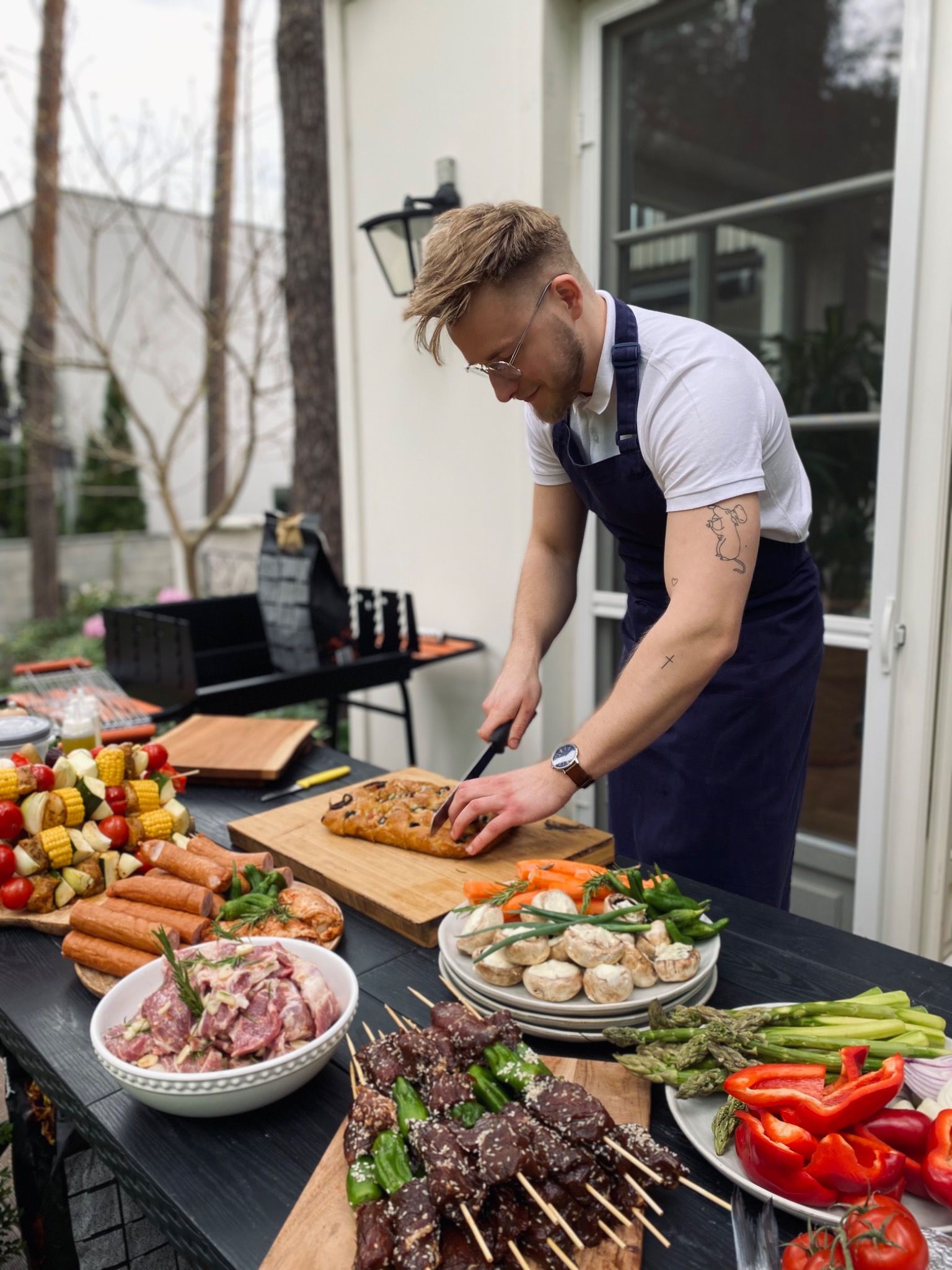 Mężczyzna w granatowym fartuchu kroi focaccię na desce, przygotowując grillowane potrawy na zewnątrz, widoczne szaszłyki, kiełbaski i warzywa.