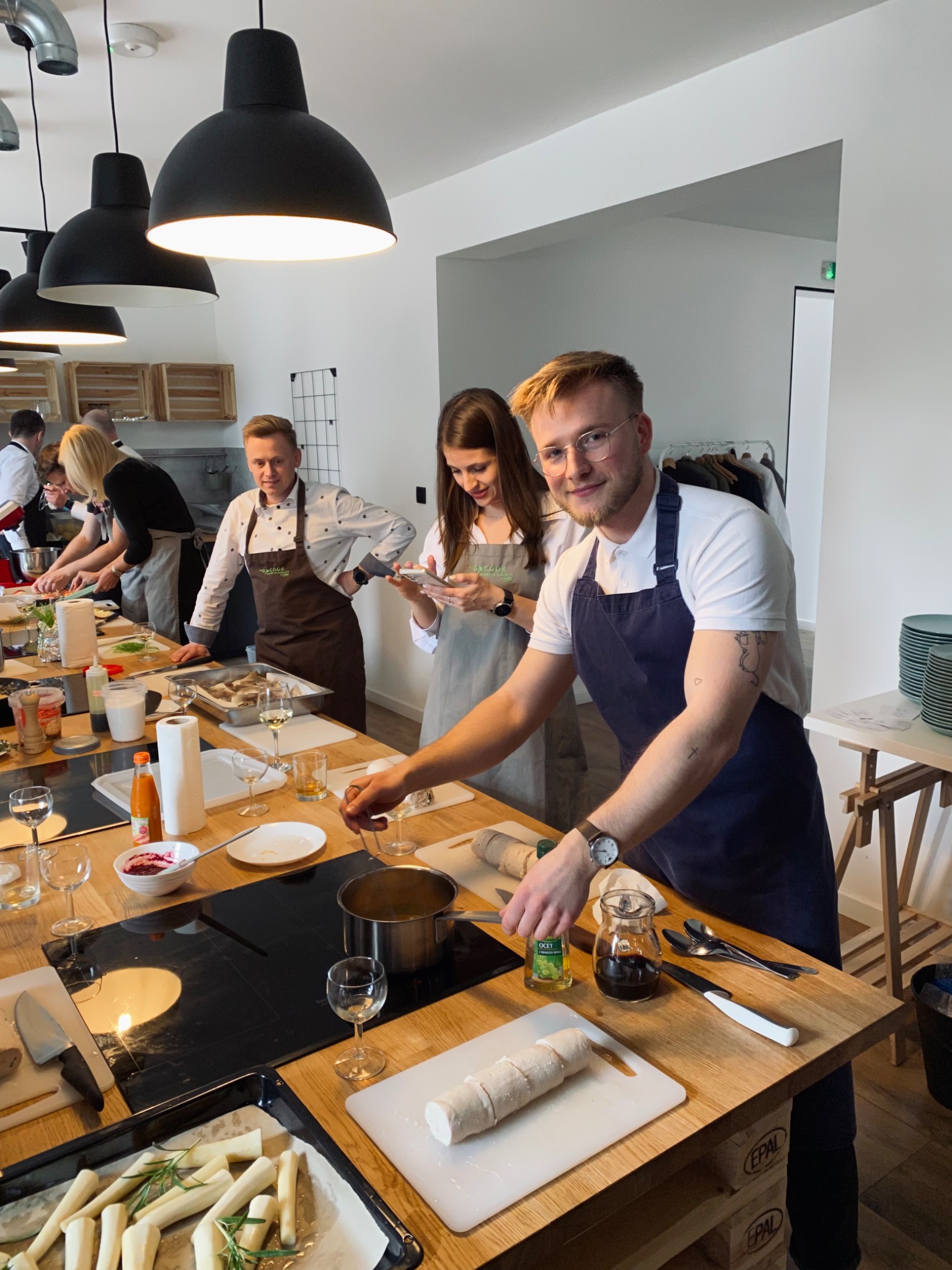 Warsztaty kulinarne: uśmiechnięty mężczyzna w fartuchu miesza składniki w garnku na indukcyjnej płycie, w tle inni uczestnicy przygotowują posiłek, widoczne składniki, przyprawy i naczynia.