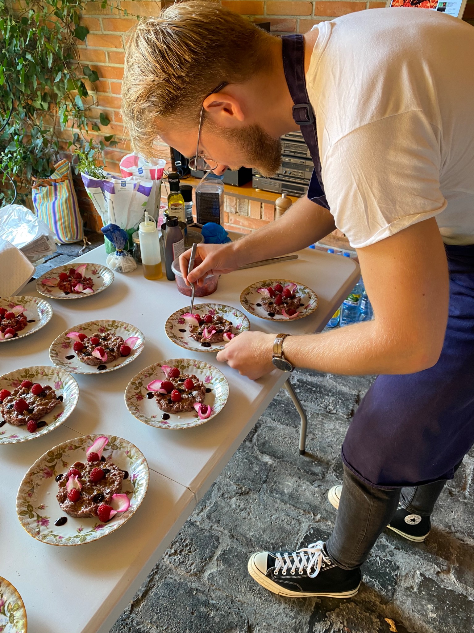 Kucharz w granatowym fartuchu dekoruje deser z malinami i płatkami róży na talerzach w kwiaty, używając precyzyjnie pędzelka.
