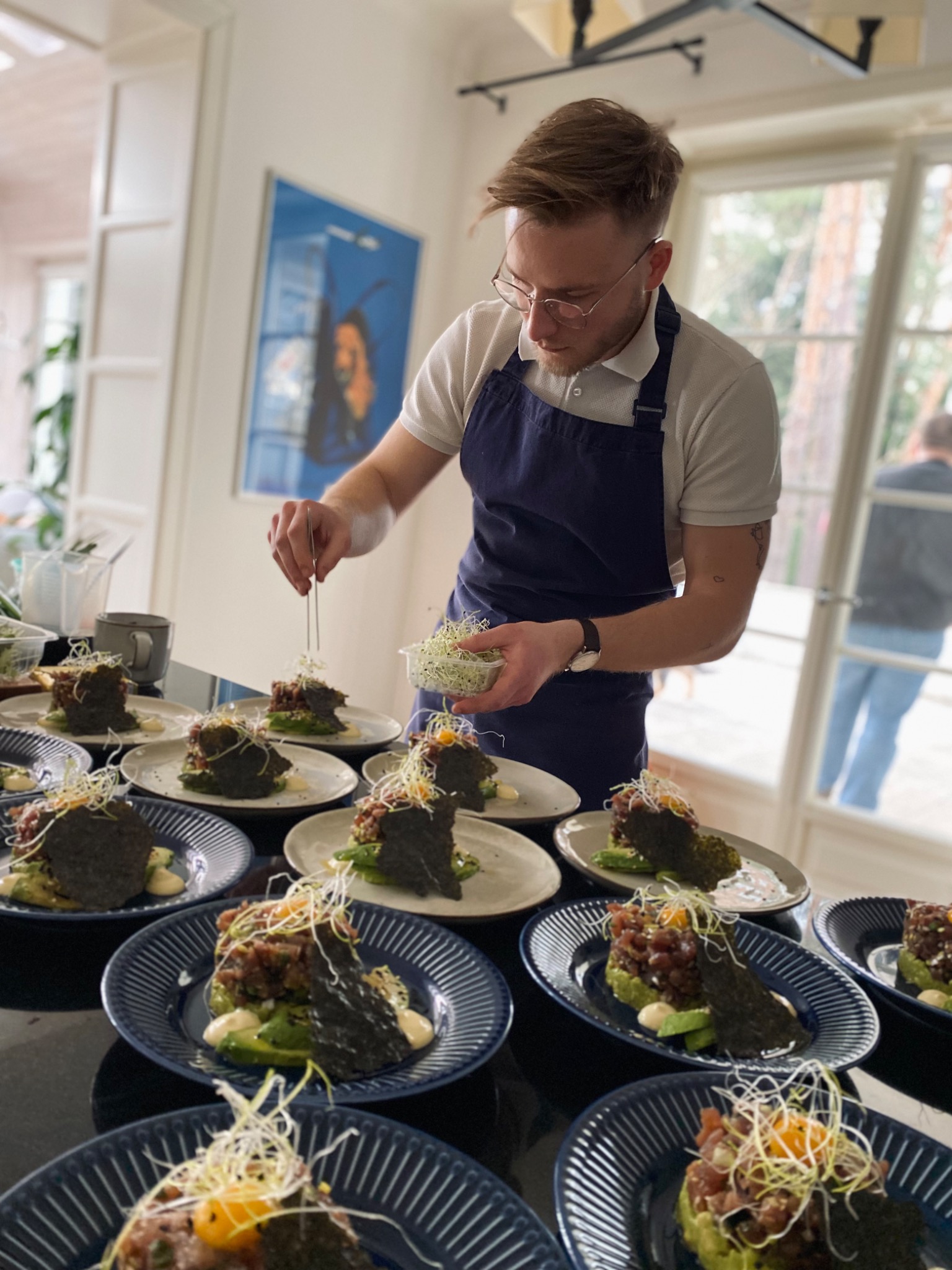 Kucharz w okularach i granatowym fartuchu dekoruje mikro-ziołami talerze z tatarem z tuńczyka na awokado z nori, przygotowane do serwowania.