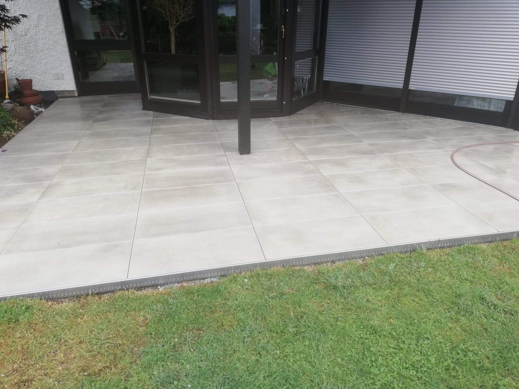 Jasnoszare płytki ceramiczne ułożone na tarasie przylegającym do domu z ciemnymi, przeszklonymi drzwiami przesuwnymi i roletami zewnętrznymi, widoczne trawnik i system odprowadzania wody.