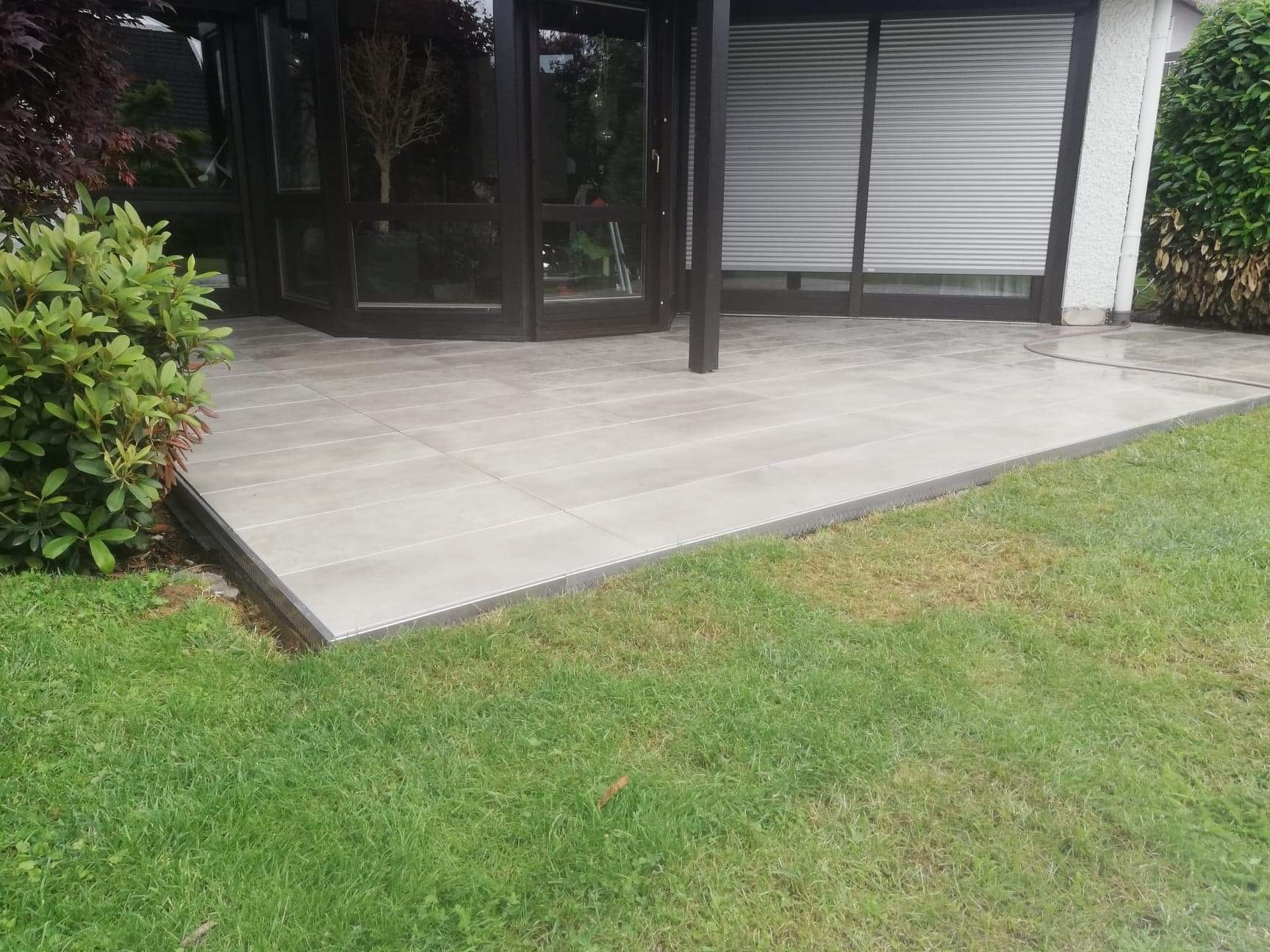 Nowoczesny taras wyłożony szarymi płytkami ceramicznymi z metalowym obrzeżem, przylegający do domu z przeszkloną werandą i roletami zewnętrznymi, otoczony trawnikiem i zielenią.