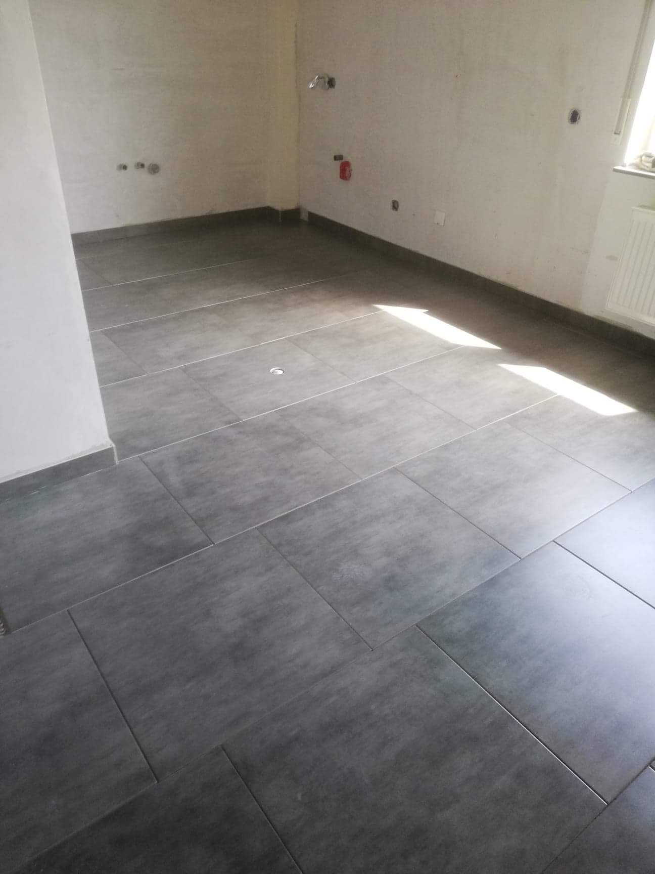 Ułożona szara, matowa podłoga z płytek ceramicznych w pustym pomieszczeniu w Częstochowie, widoczne instalacje na ścianach i fragment okna z grzejnikiem.