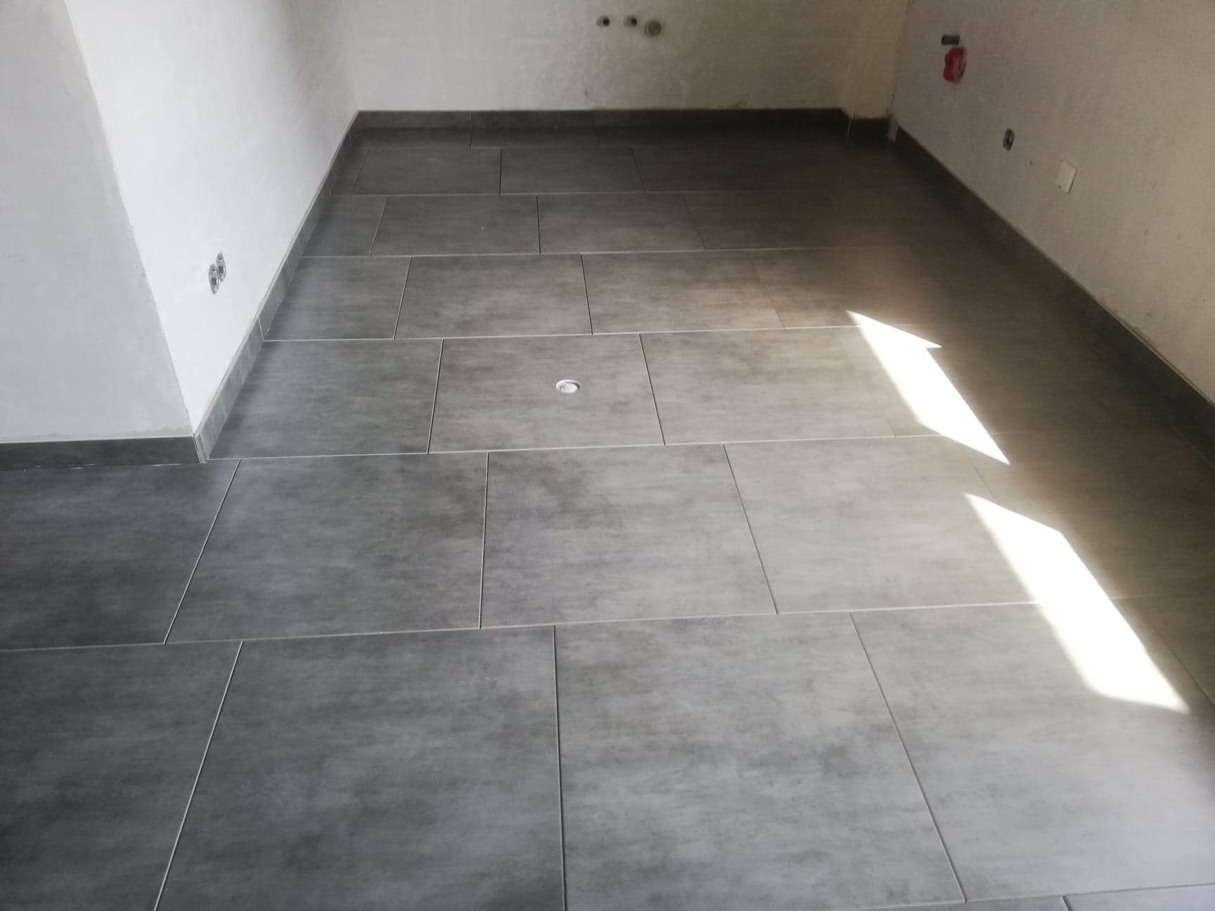 Ułożone szare płytki podłogowe w pomieszczeniu w trakcie prac wykończeniowych, widoczne ślady po instalacjach na ścianie i intensywne światło słoneczne wpadające przez okno.